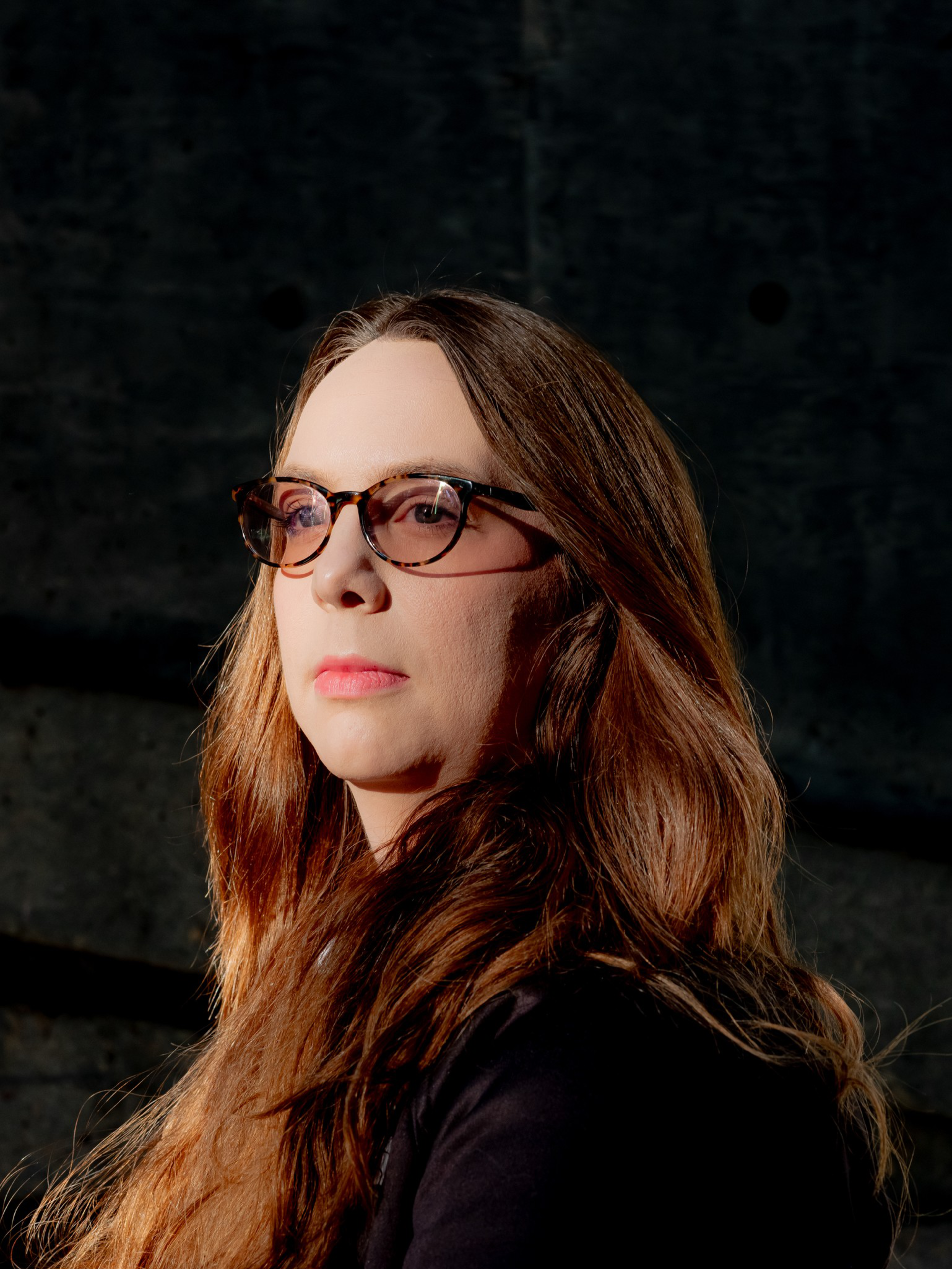 A woman with long brown hair and glasses looks thoughtfully into the distance against a dark, blurred background.
