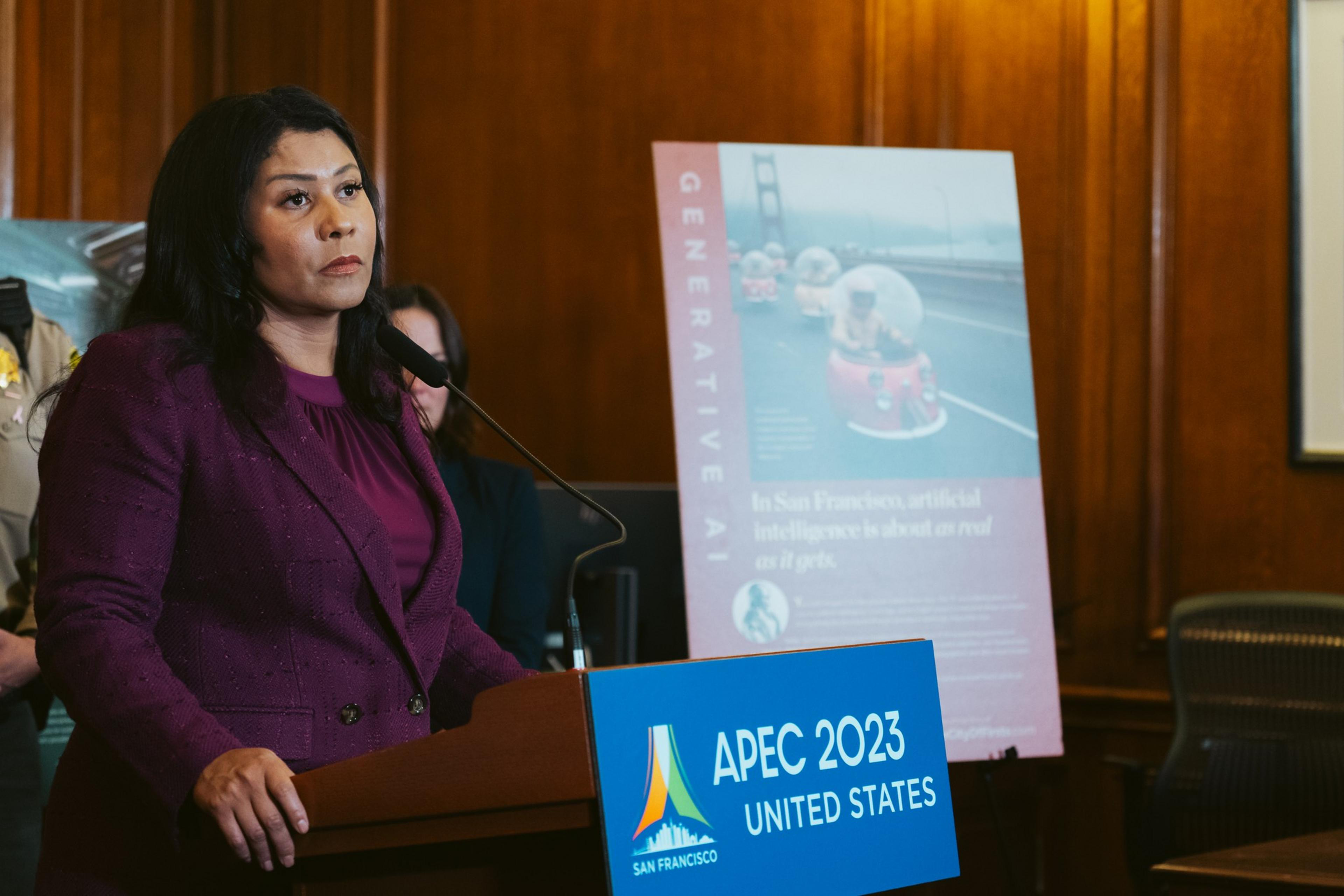 A woman in a magenta jacket speaks at a podium with &quot;APEC 2023 UNITED STATES&quot; displayed, with a poster about AI and San Francisco's Golden Gate Bridge in the background.
