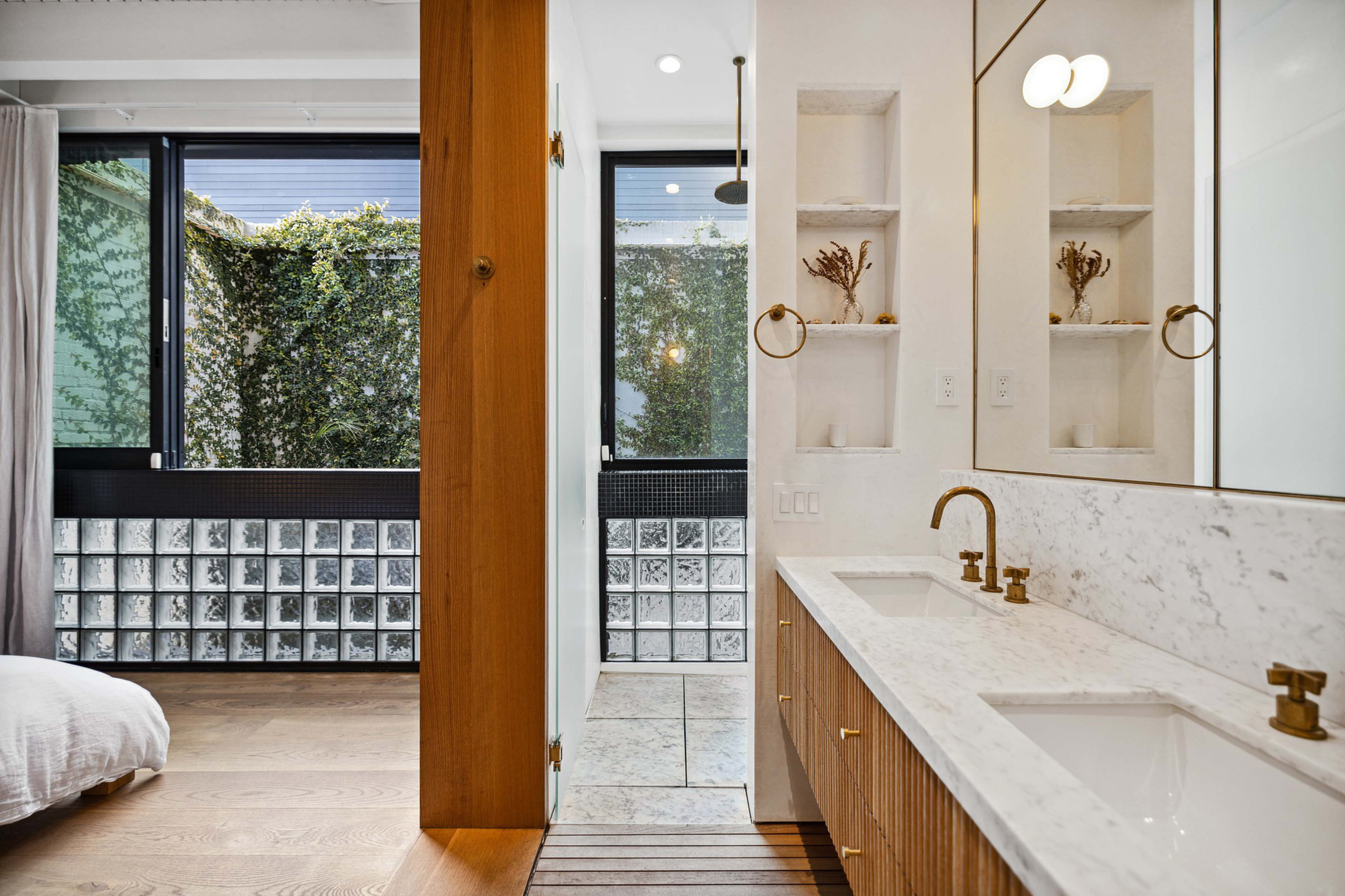A modern bathroom with a double sink vanity, gold fixtures, a large mirror, built-in shelves with dried plants, and a glass-enclosed shower; a bedroom is partially visible.
