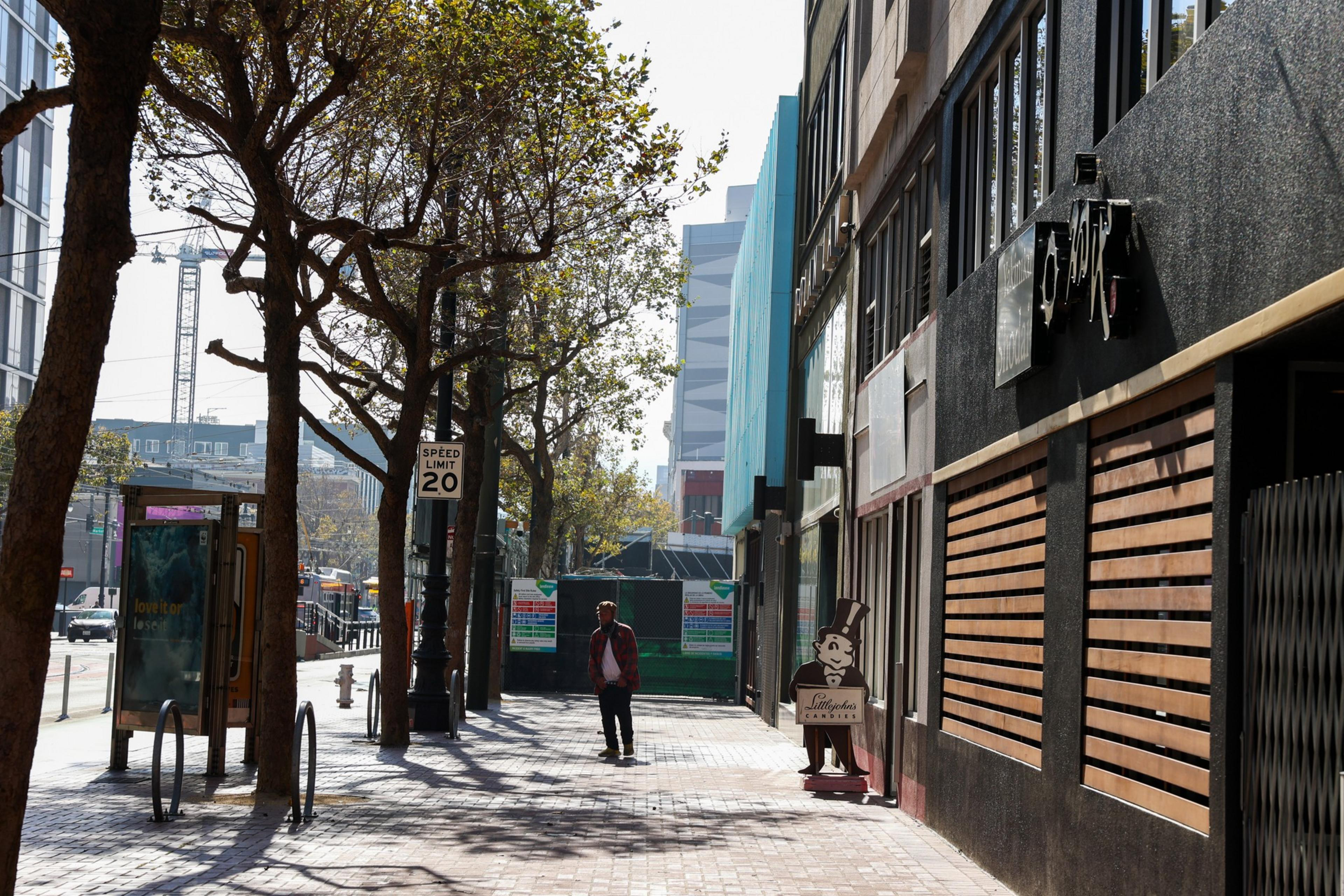 A sunny sidewalk lined with trees beside a building, with a figure walking, and a speed limit sign showing 20 mph.