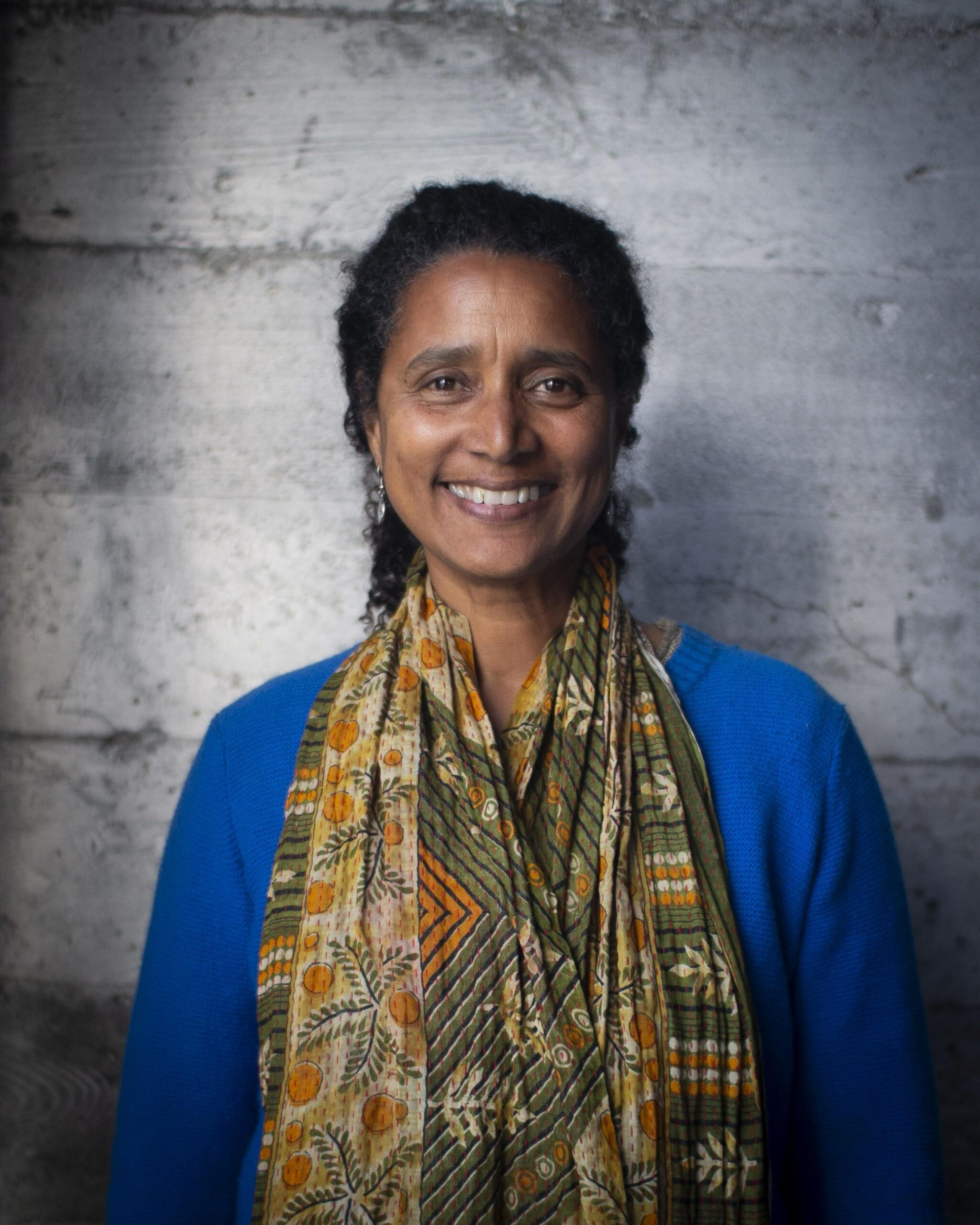 A smiling woman with curly hair wears a blue top and a patterned scarf with orange and green designs, standing in front of a textured gray wall.