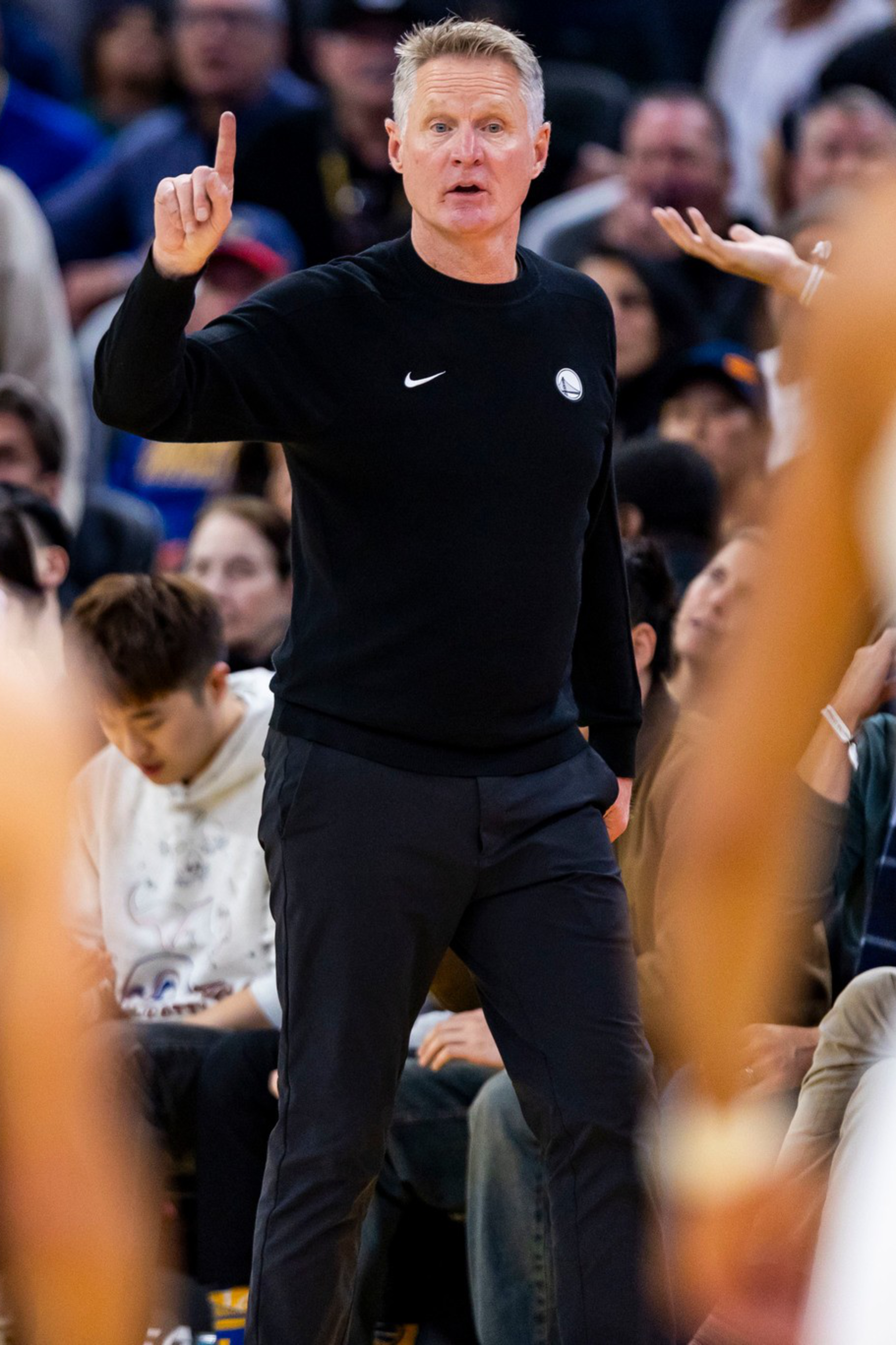 A basketball coach in black gestures with one finger raised while players in blue and white uniforms face him during a game with a crowd behind.