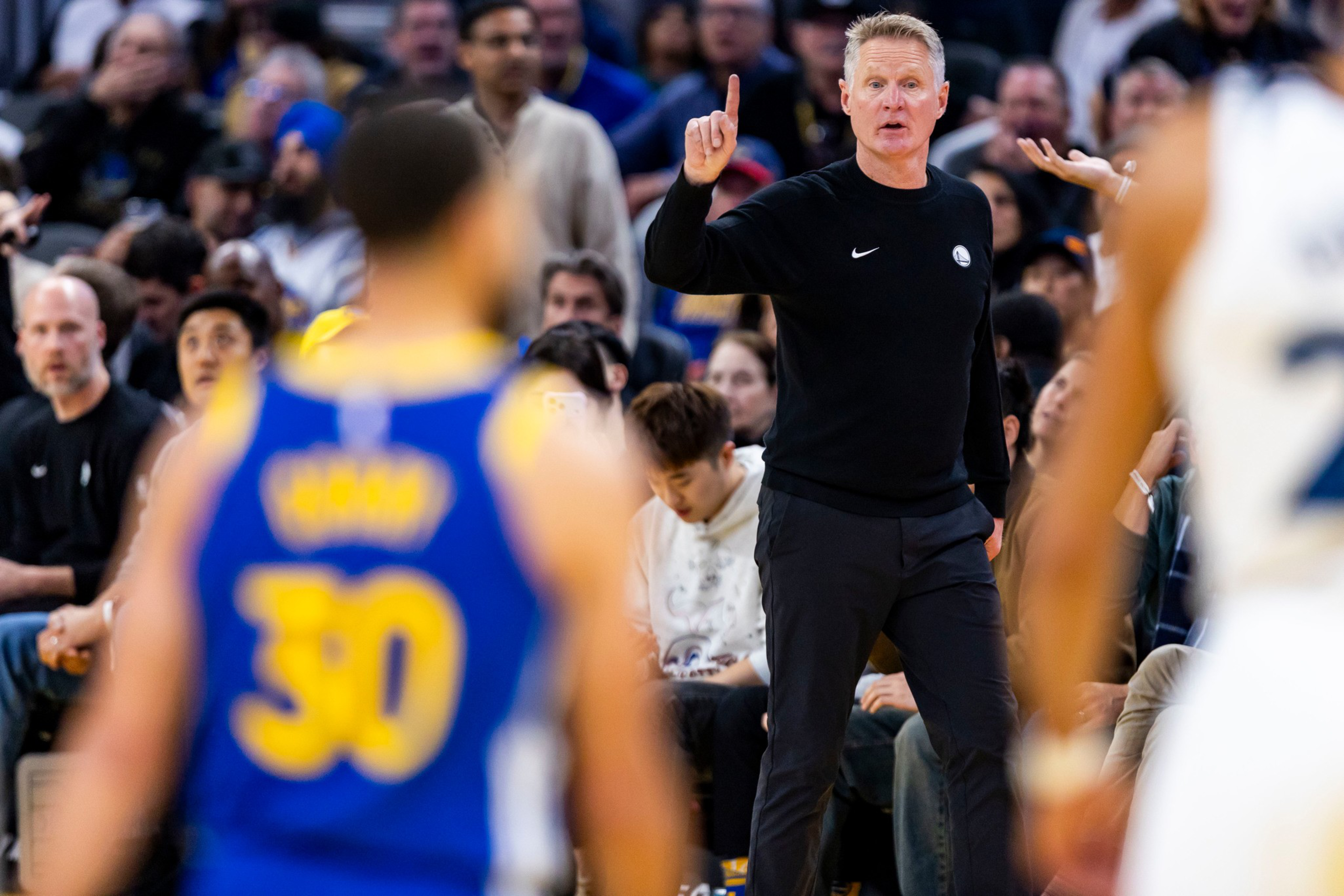 A basketball coach in black gestures with one finger raised while players in blue and white uniforms face him during a game with a crowd behind.