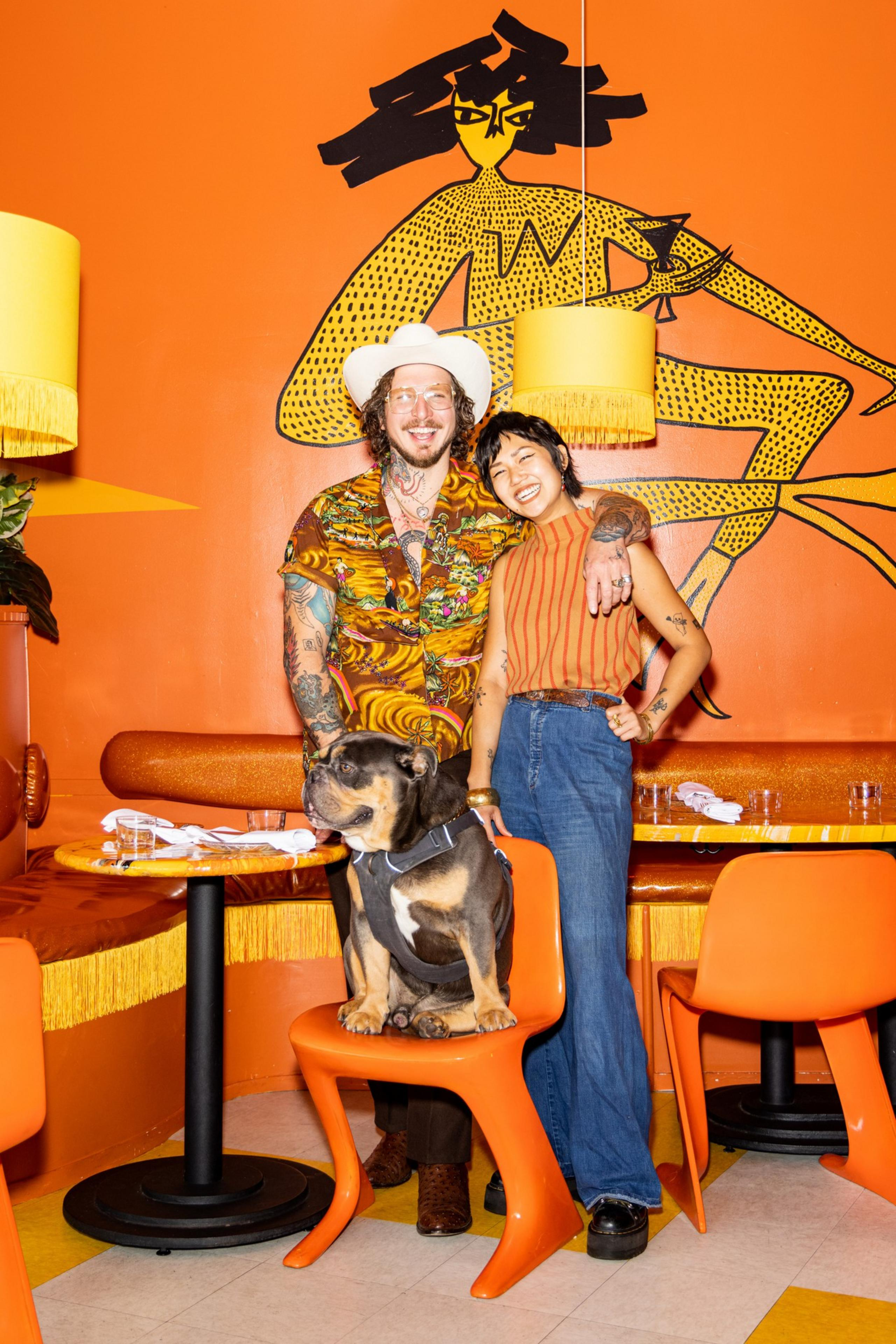 A tattooed man in a patterned shirt and white hat hugs a smiling woman in a striped top and jeans, while a bulldog sits on an orange chair.