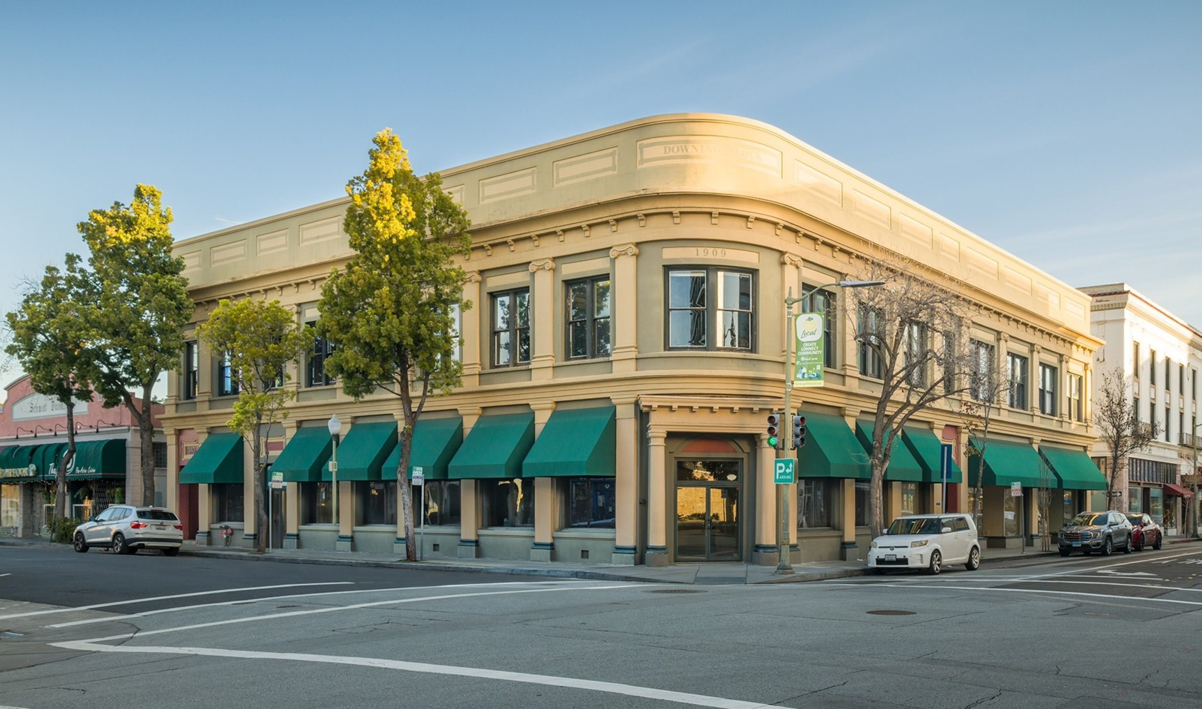 A two-story corner building with green awnings over the windows, beige walls, and several trees lining the sidewalk on a clear day.