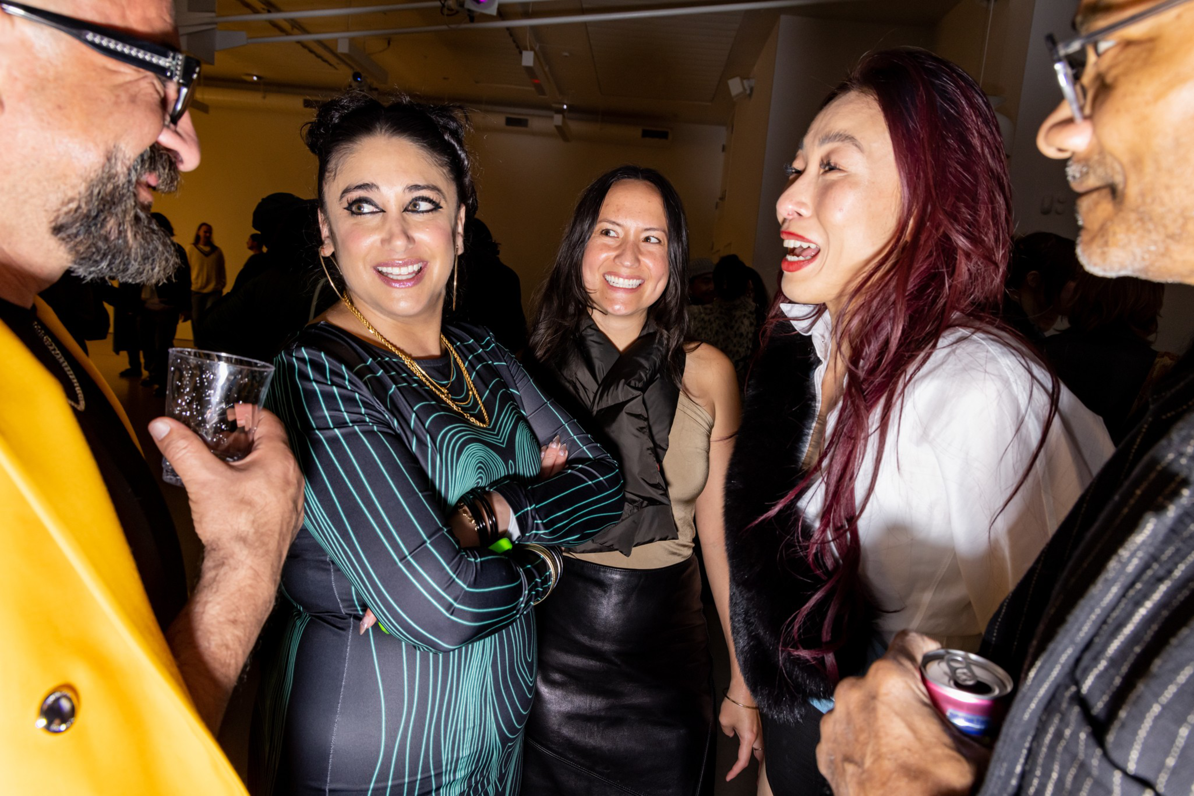 A group of people stand close together at a lively gathering, engaged in animated conversation, with some holding drinks and smiling brightly at each other.