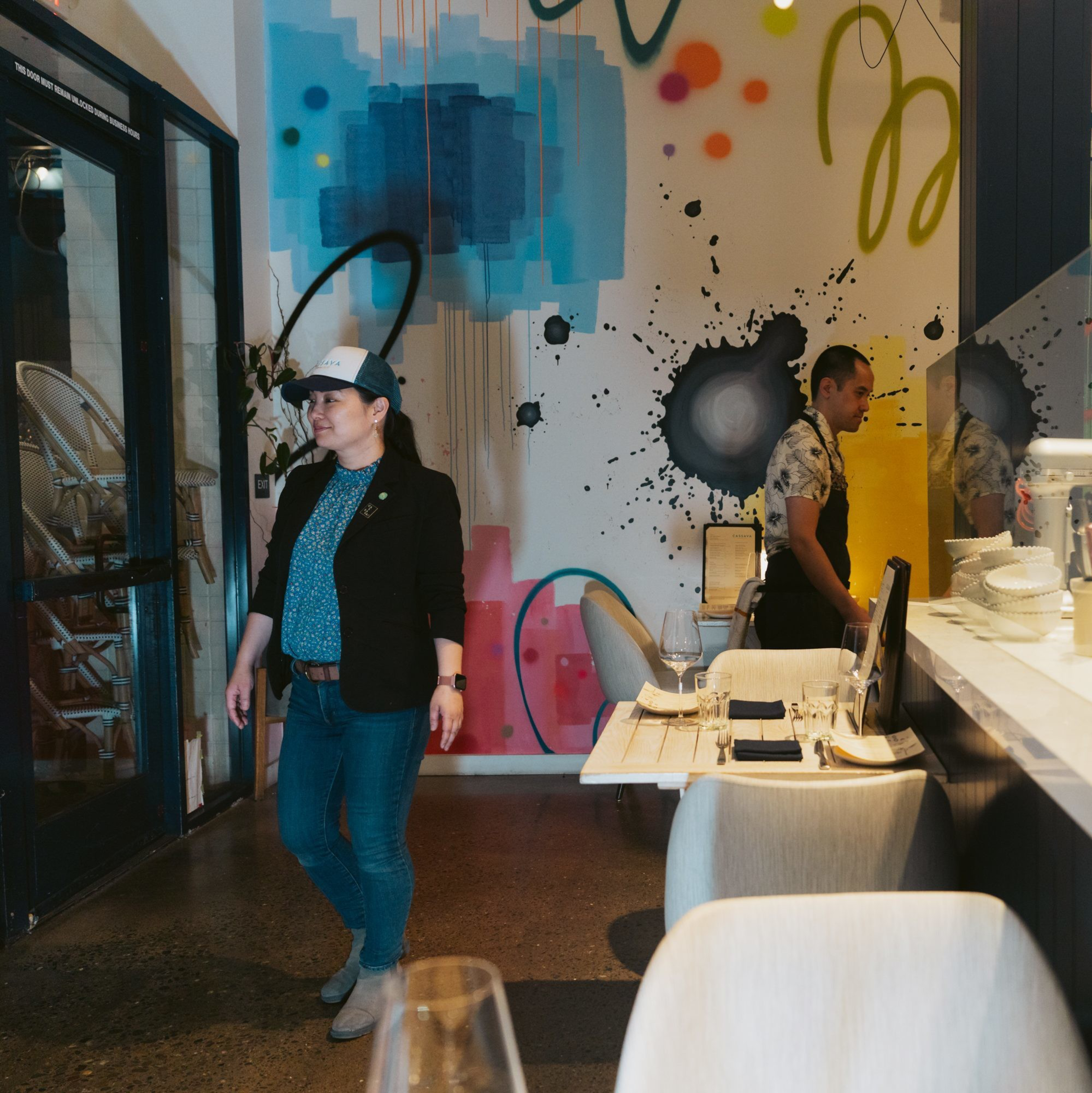A woman in casual attire walks through a stylish restaurant with colorful abstract wall art. A man is working near a counter, setting up place settings.