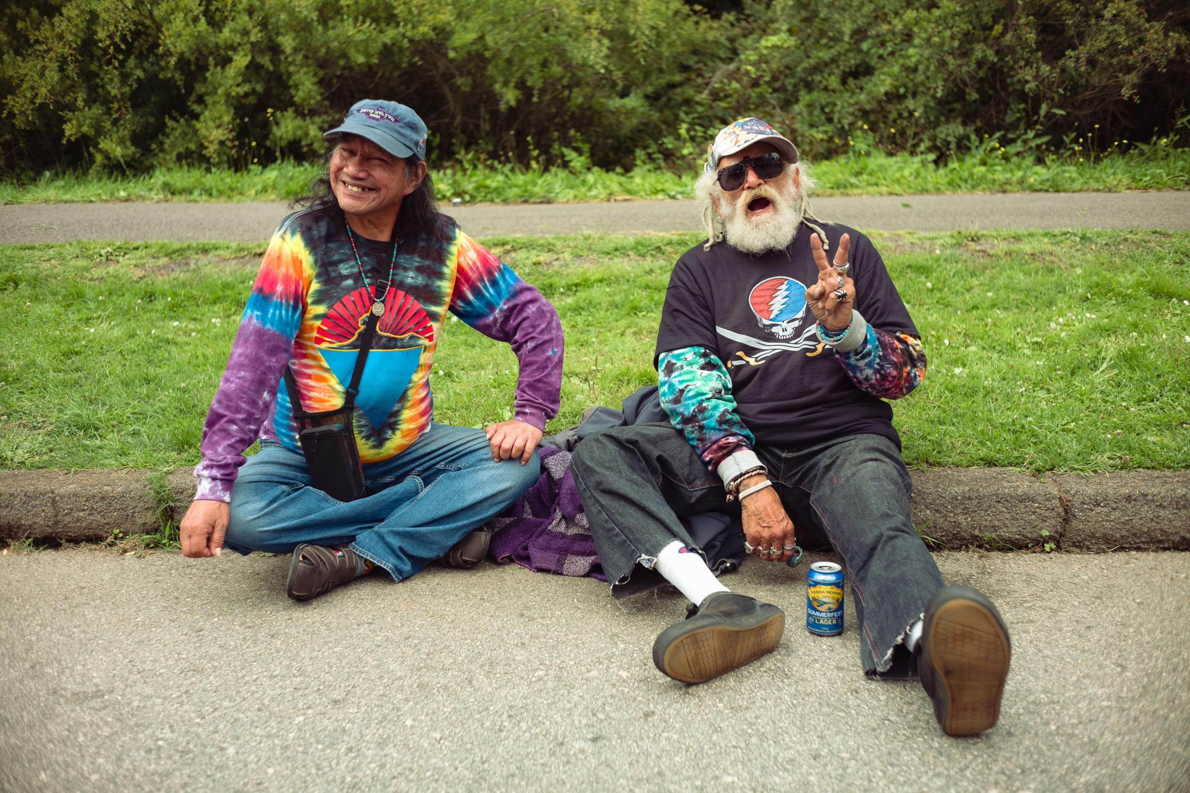 Two older men sit on the ground by grass; one wears a colorful tie-dye shirt and cap, the other sports sunglasses, a black shirt, and shows a peace sign.