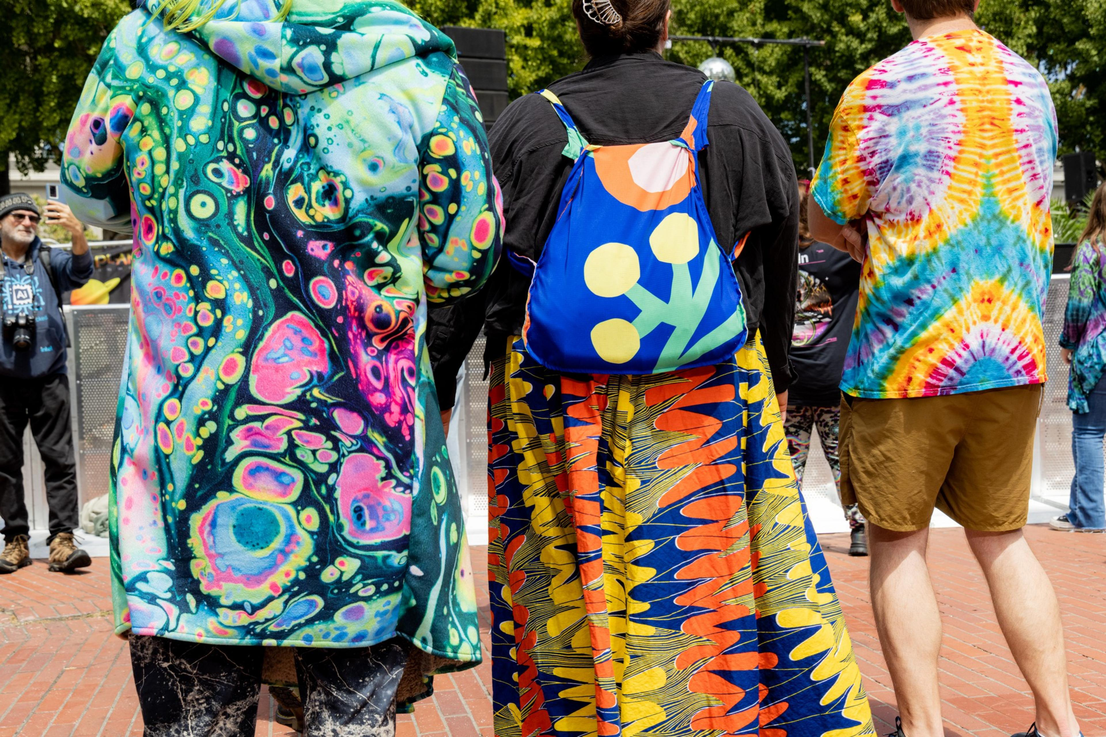 Three individuals wearing colorful, psychedelic-patterned clothing stand outside. One has a tie-dye shirt, another has a vibrant dress, and the last wears a vivid coat.