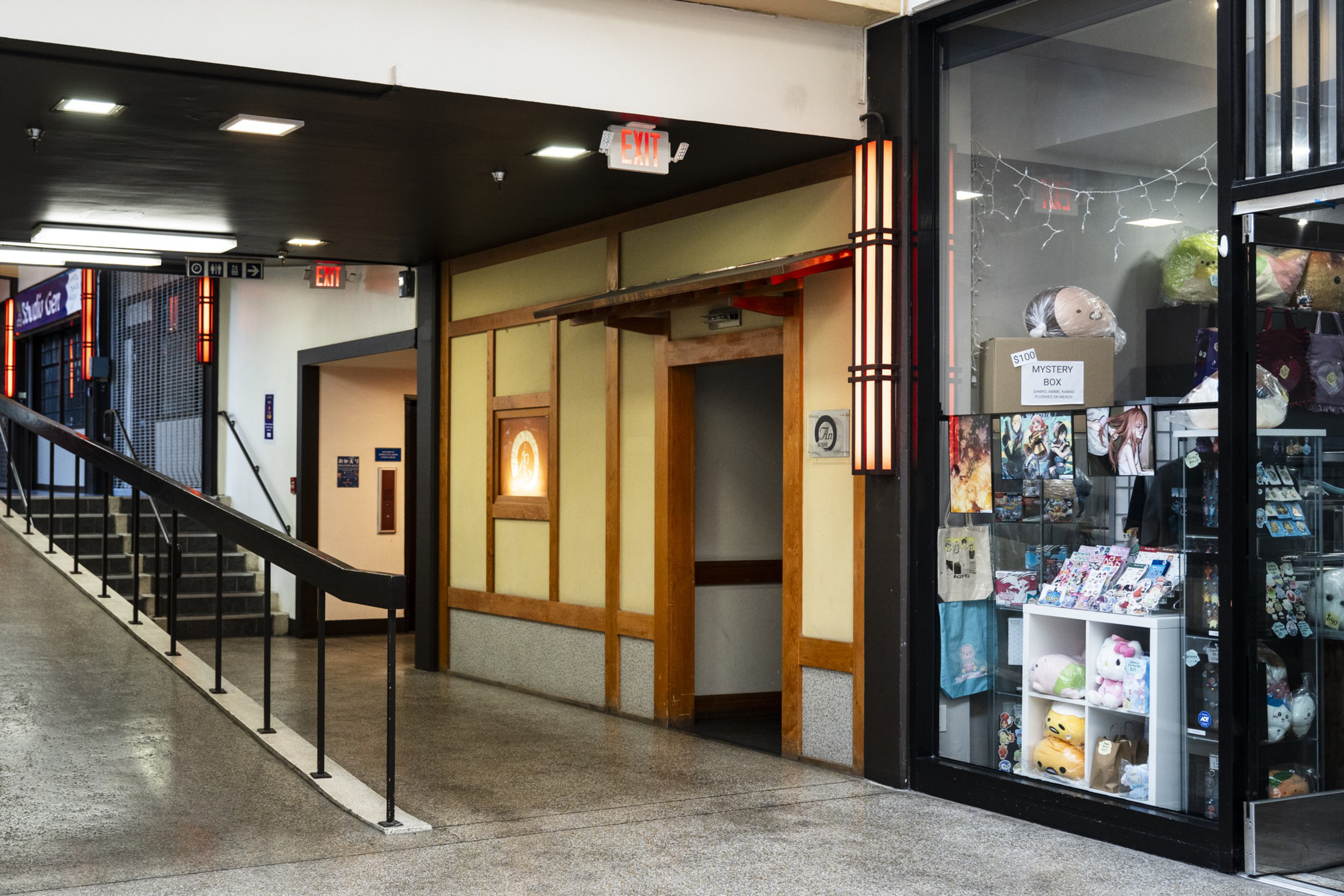 A hallway with a descending ramp and stairs on the left, a simple wooden door in the center, and a glass storefront displaying plush toys and art on the right.
