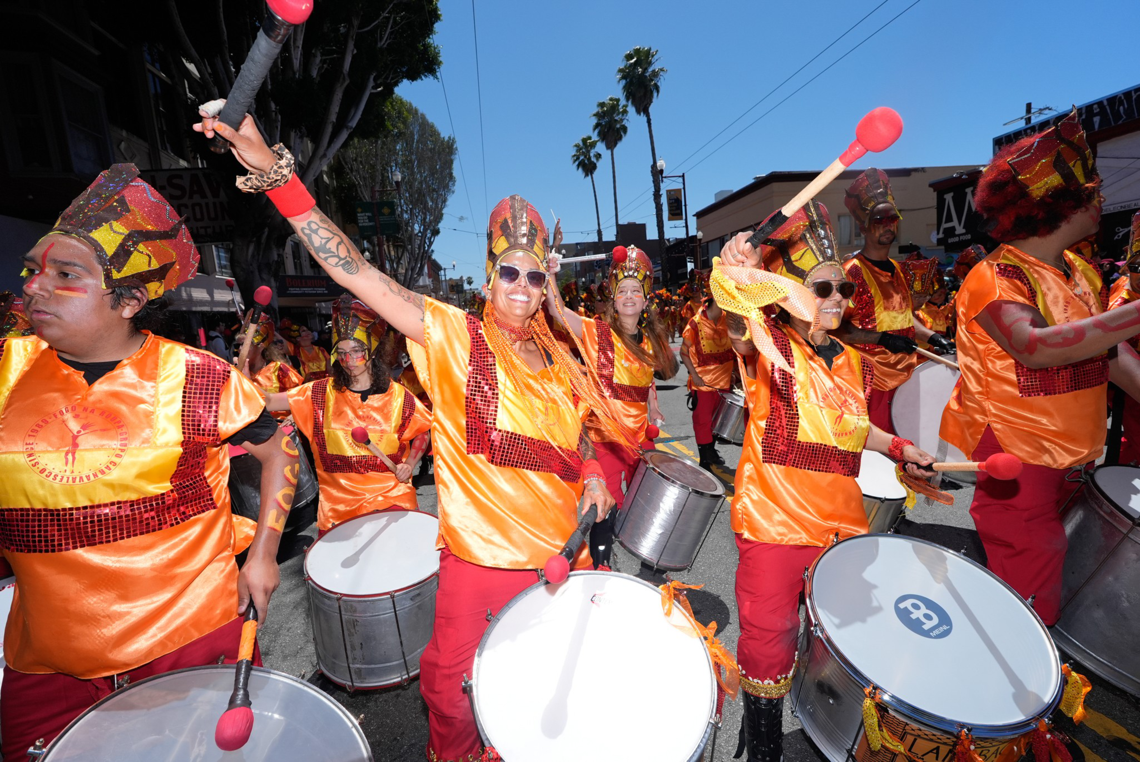 A drum troupe wearing brightly colored costumes hold mallets high over their heads against a backdrop of palm trees and sunlit buildings.