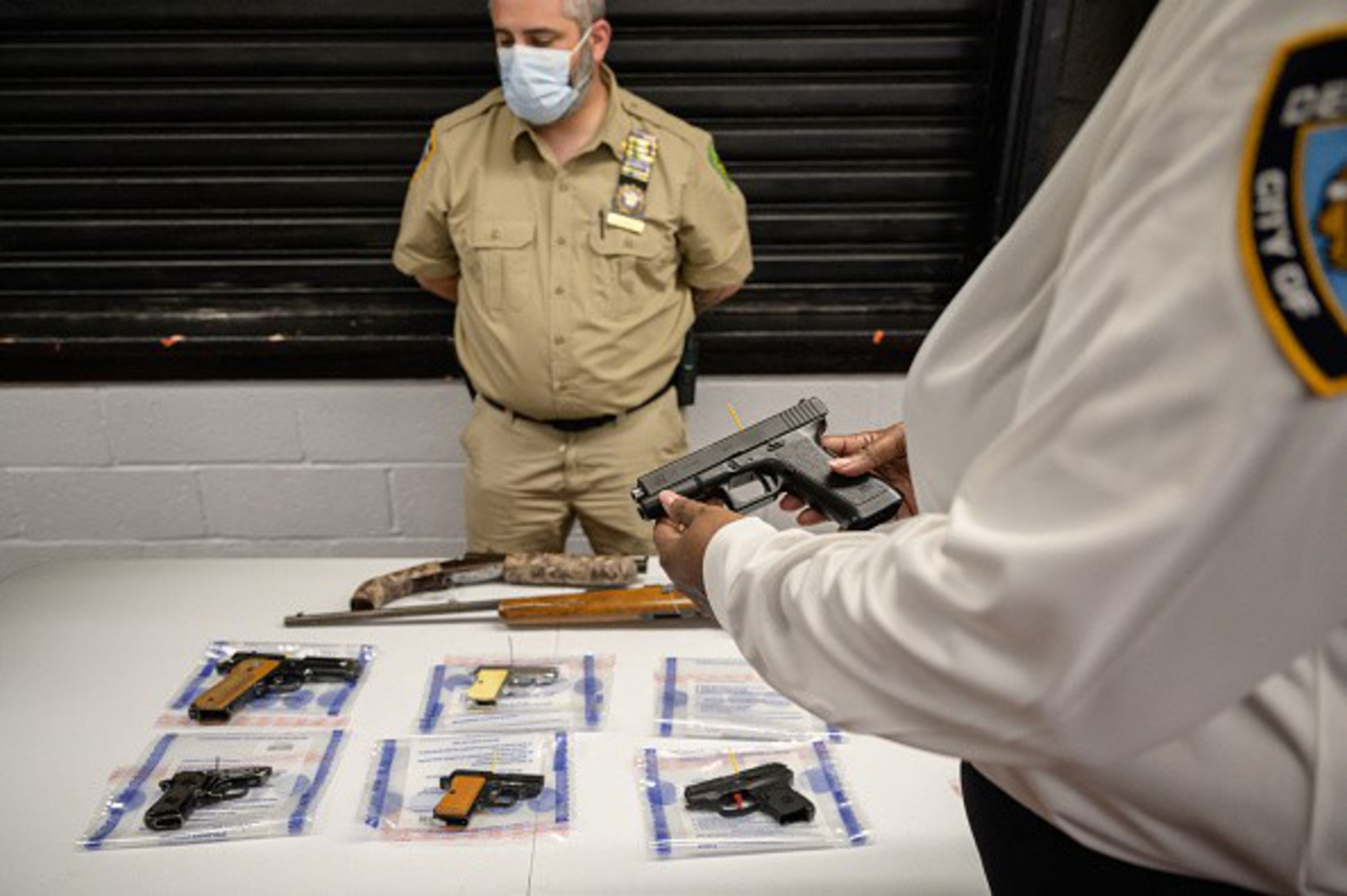 A masked officer in beige uniform stands behind a table with several firearms in evidence bags, while another officer in white holds a pistol.