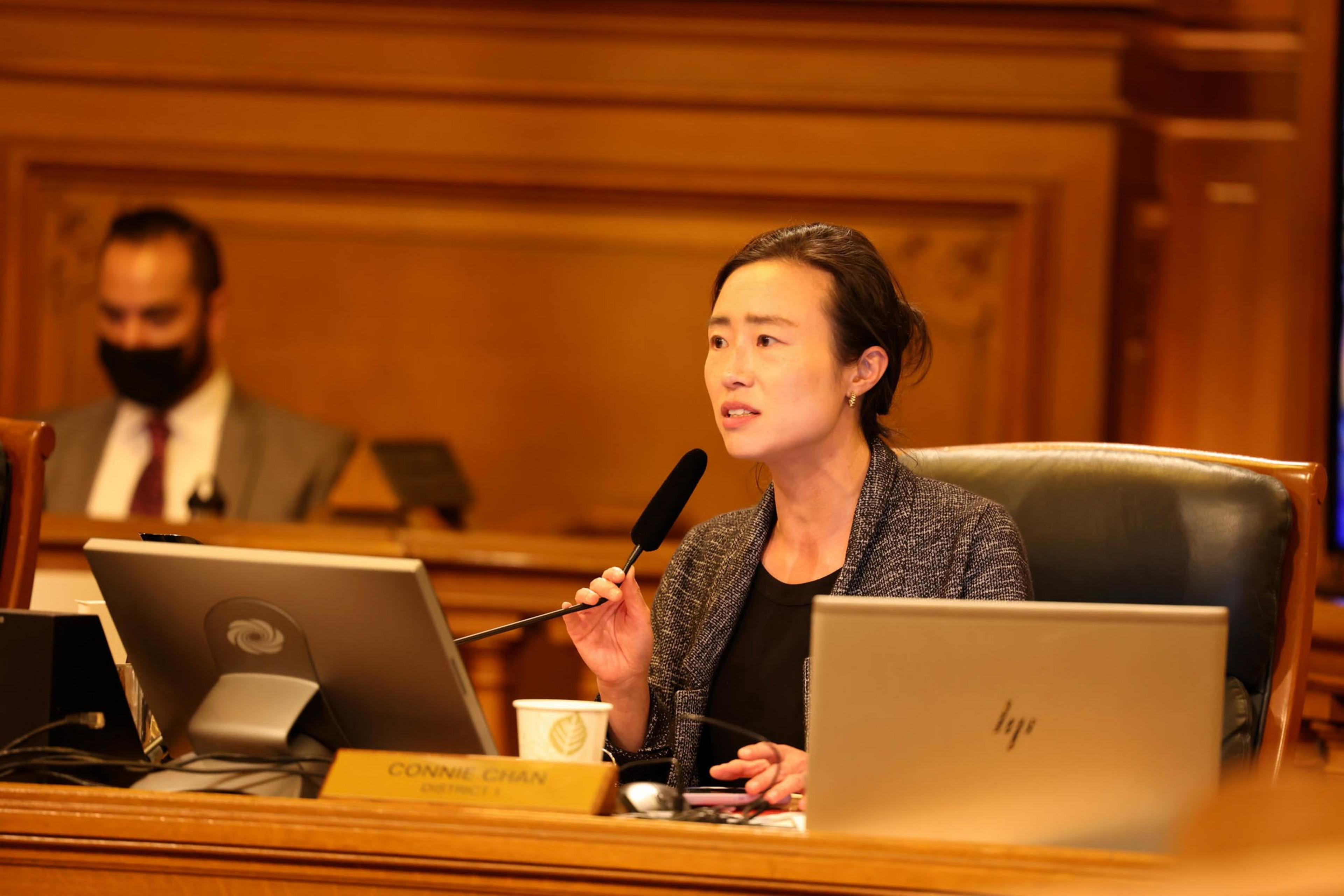 A woman speaks into a microphone during a meeting, seated at a desk with a nameplate reading "Connie Chan," surrounded by monitors and a laptop.
