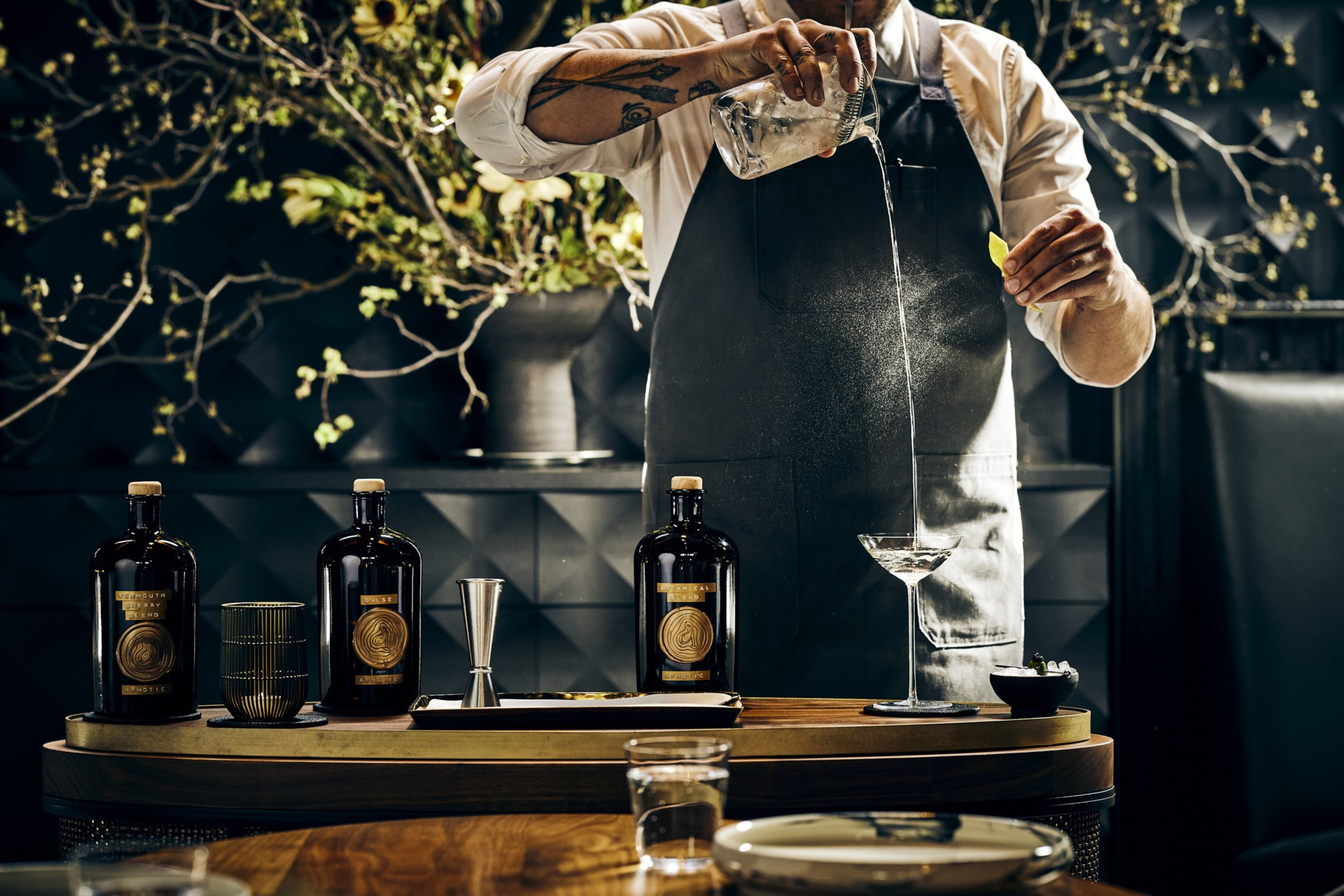 A bartender in a black apron pours a drink from a glass into a cocktail glass while holding a lemon wedge, with three dark bottles and bar tools on the counter.