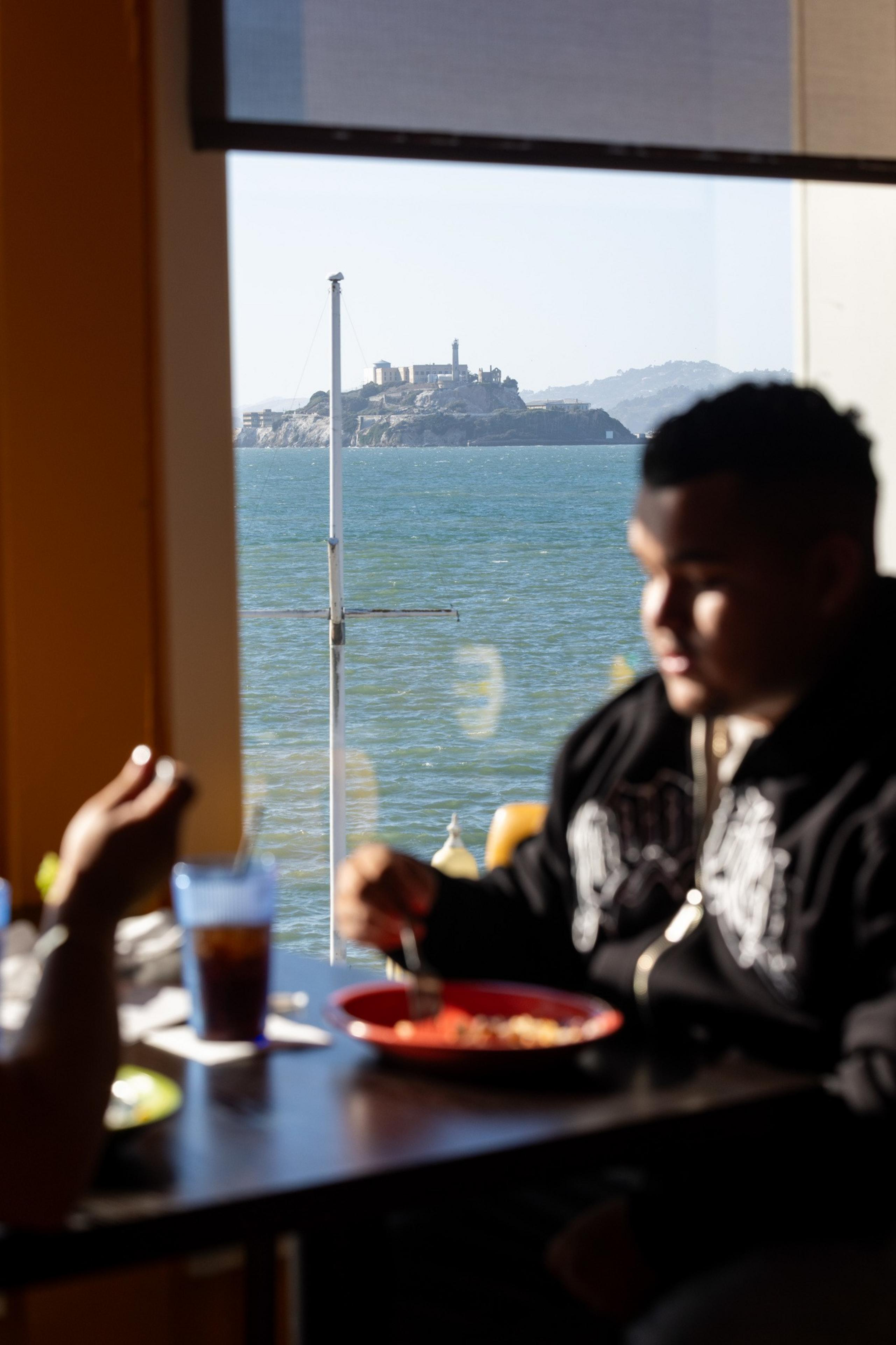 A person eats at a table by a window overlooking blue water with an island and lighthouse in the distance.