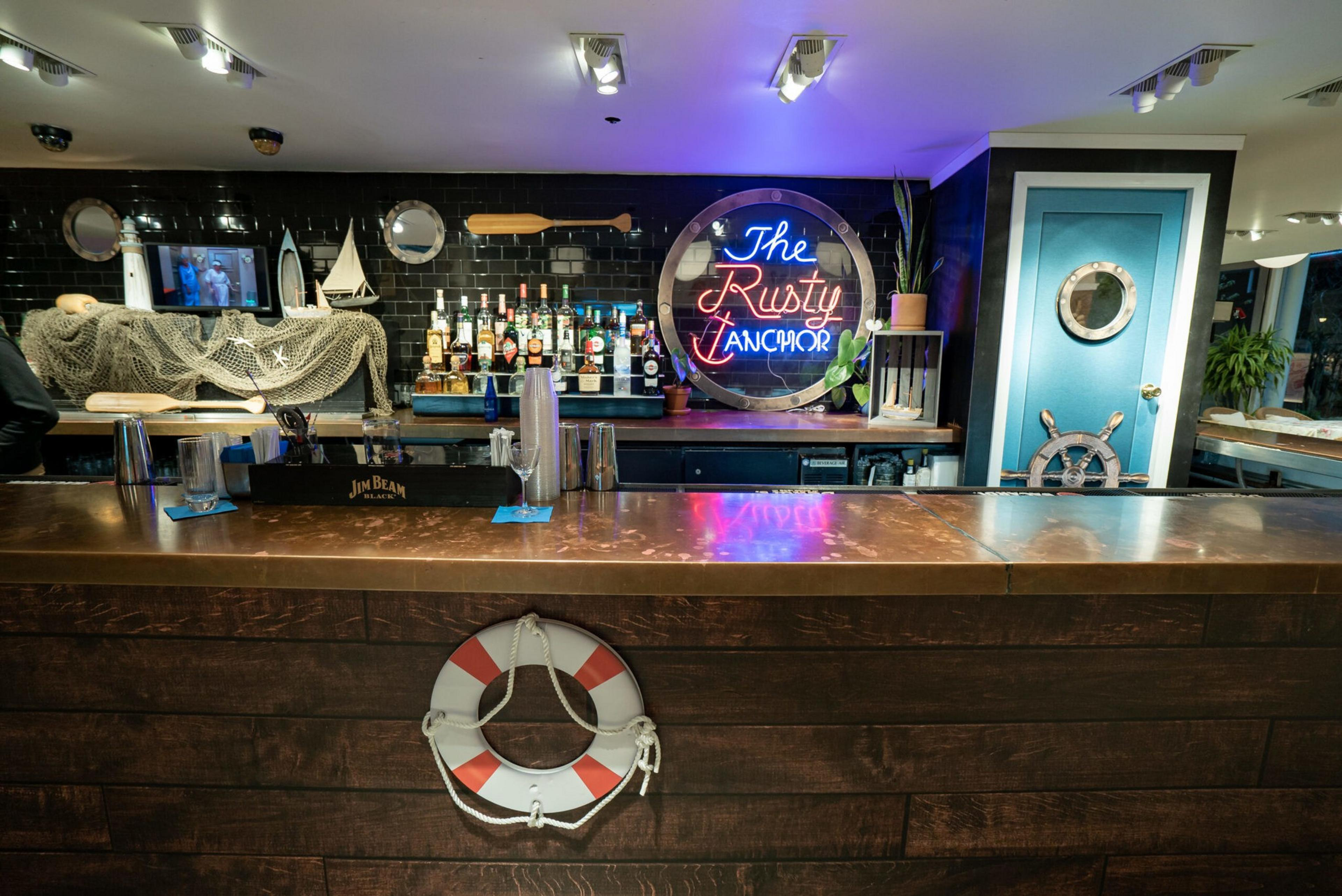 A nautical-themed bar with a lifebuoy on the front counter, a neon sign reading "The Rusty Anchor," liquor bottles, and maritime decor on black tiled walls.