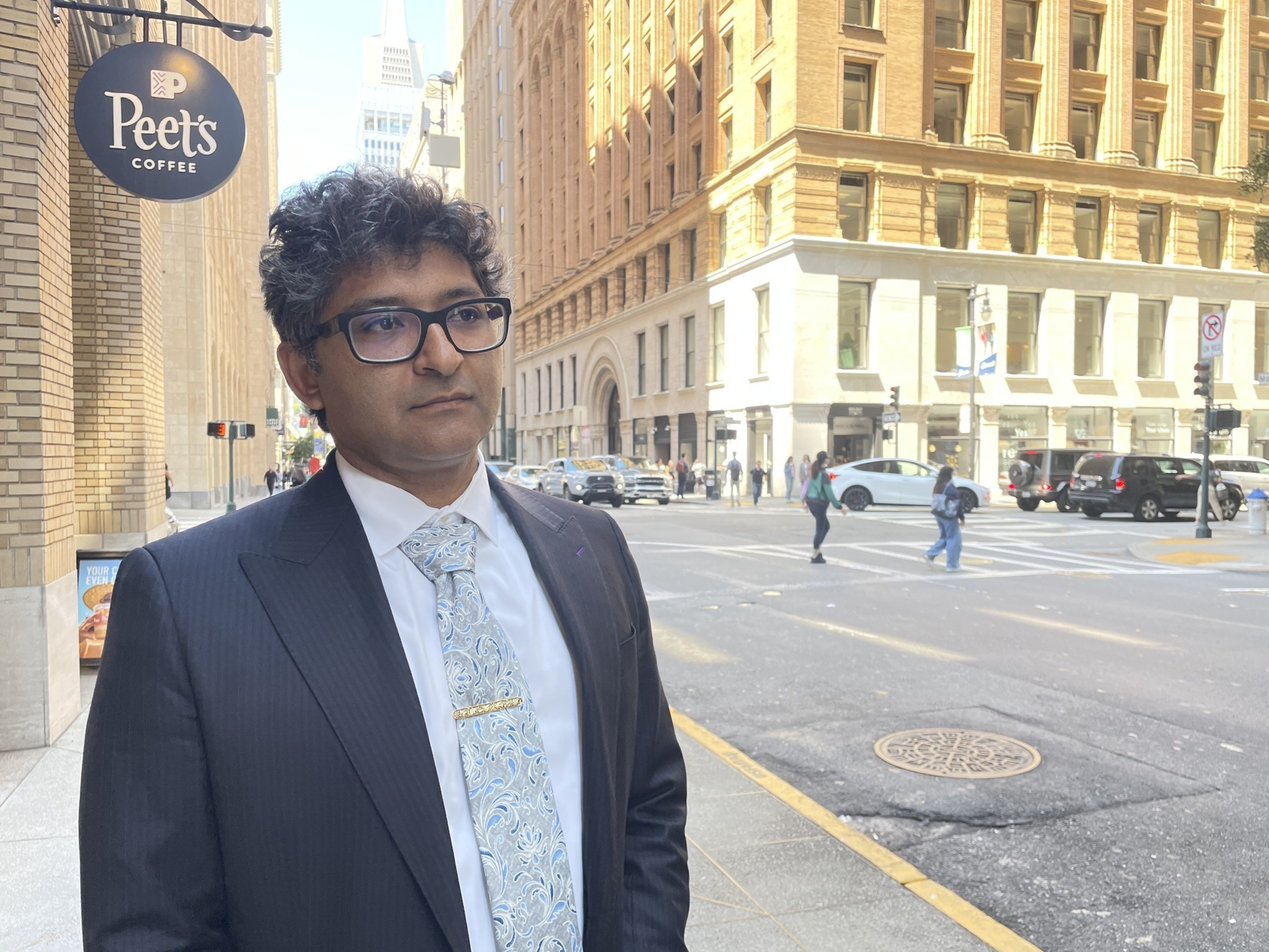A person in a suit stands on a city street near a Peet's Coffee sign. Tall buildings and pedestrians fill the bustling background.