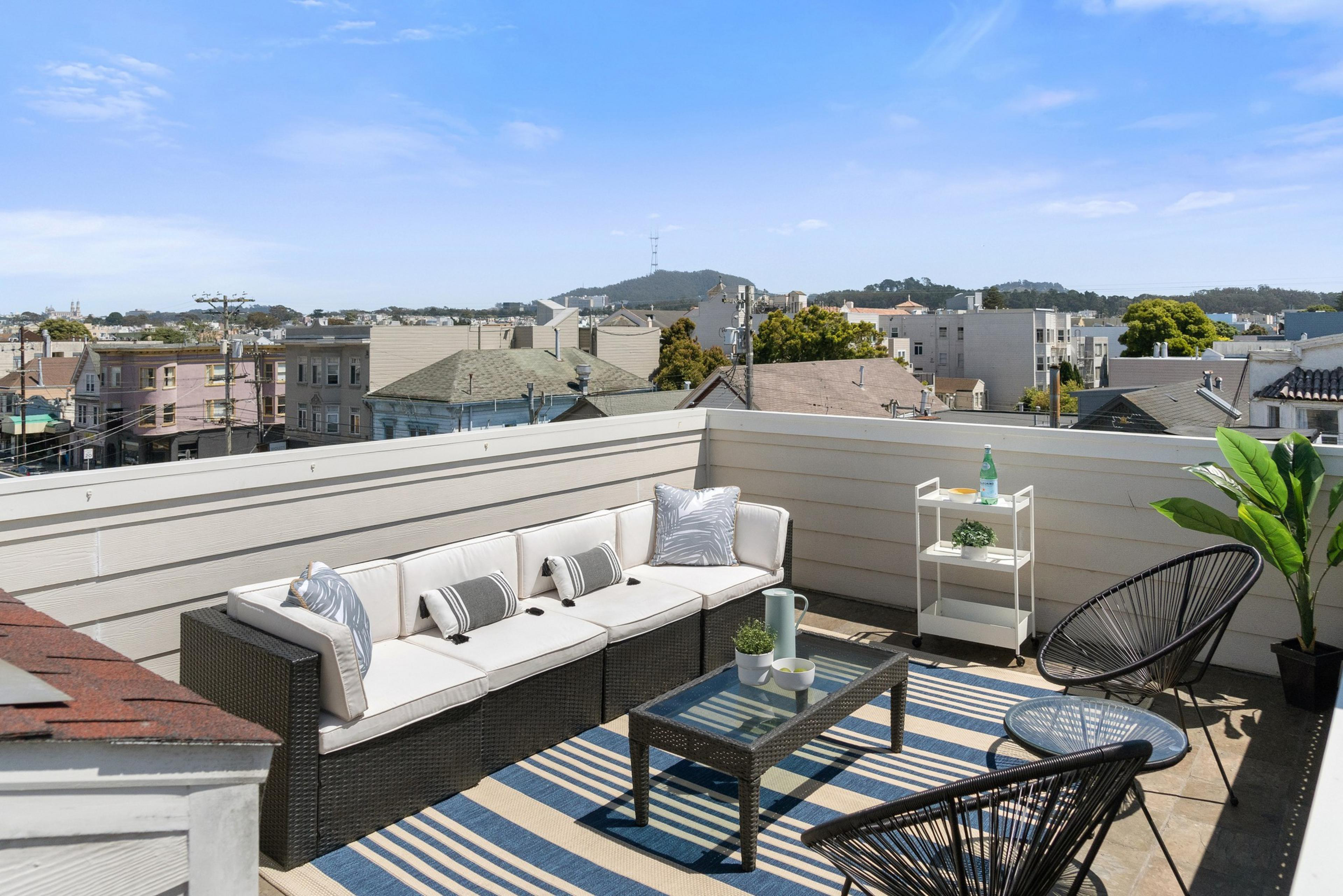 The image showcases a rooftop patio with a white sectional sofa, two black chairs, a glass coffee table, plants, and a view of a neighborhood and distant hills.