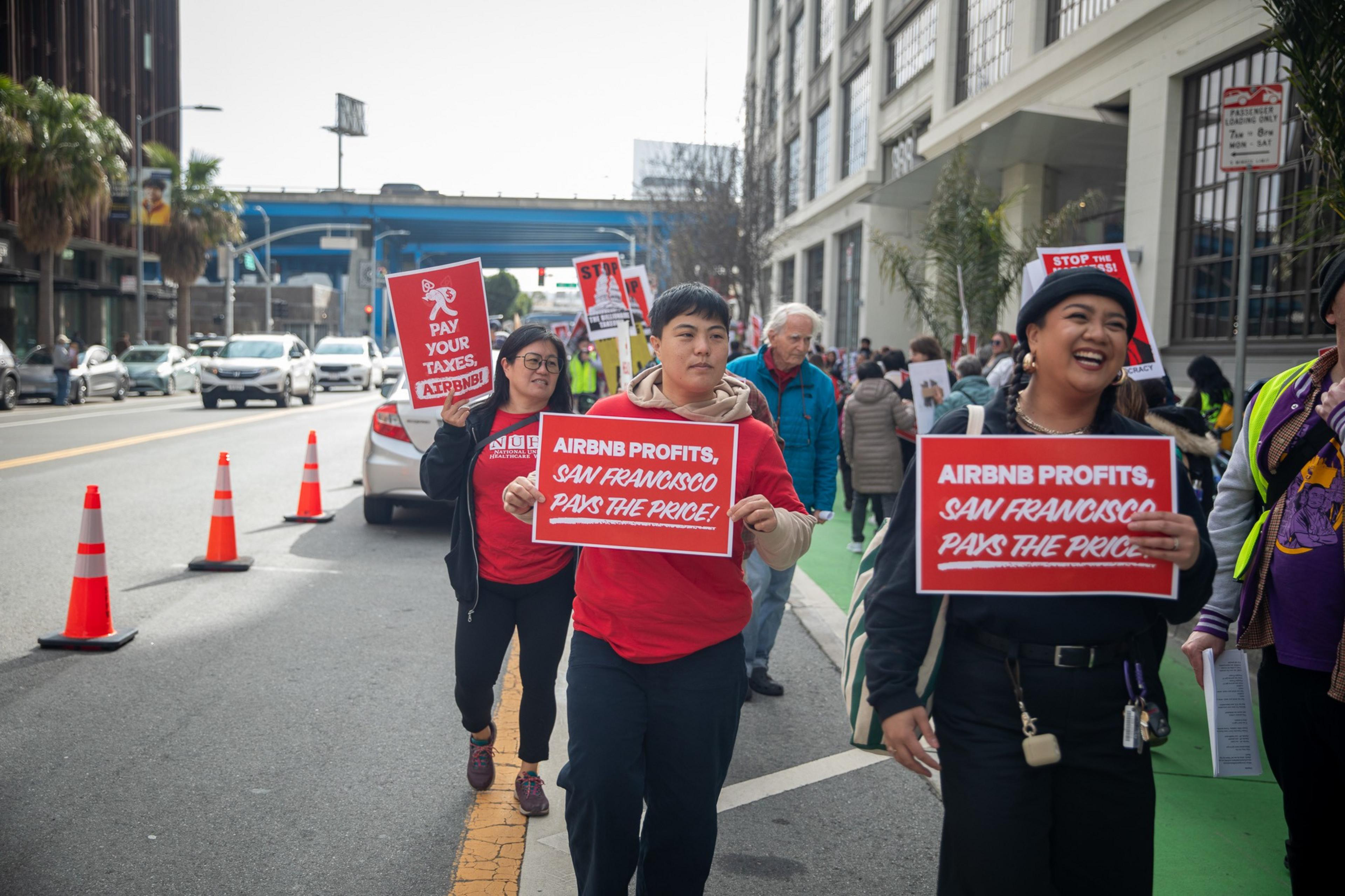 People are marching on a city street holding signs that read, &quot;AIRBNB PROFITS, SAN FRANCISCO PAYS THE PRICE!&quot; and &quot;PAY YOUR TAXES, AIRBNB.&quot;