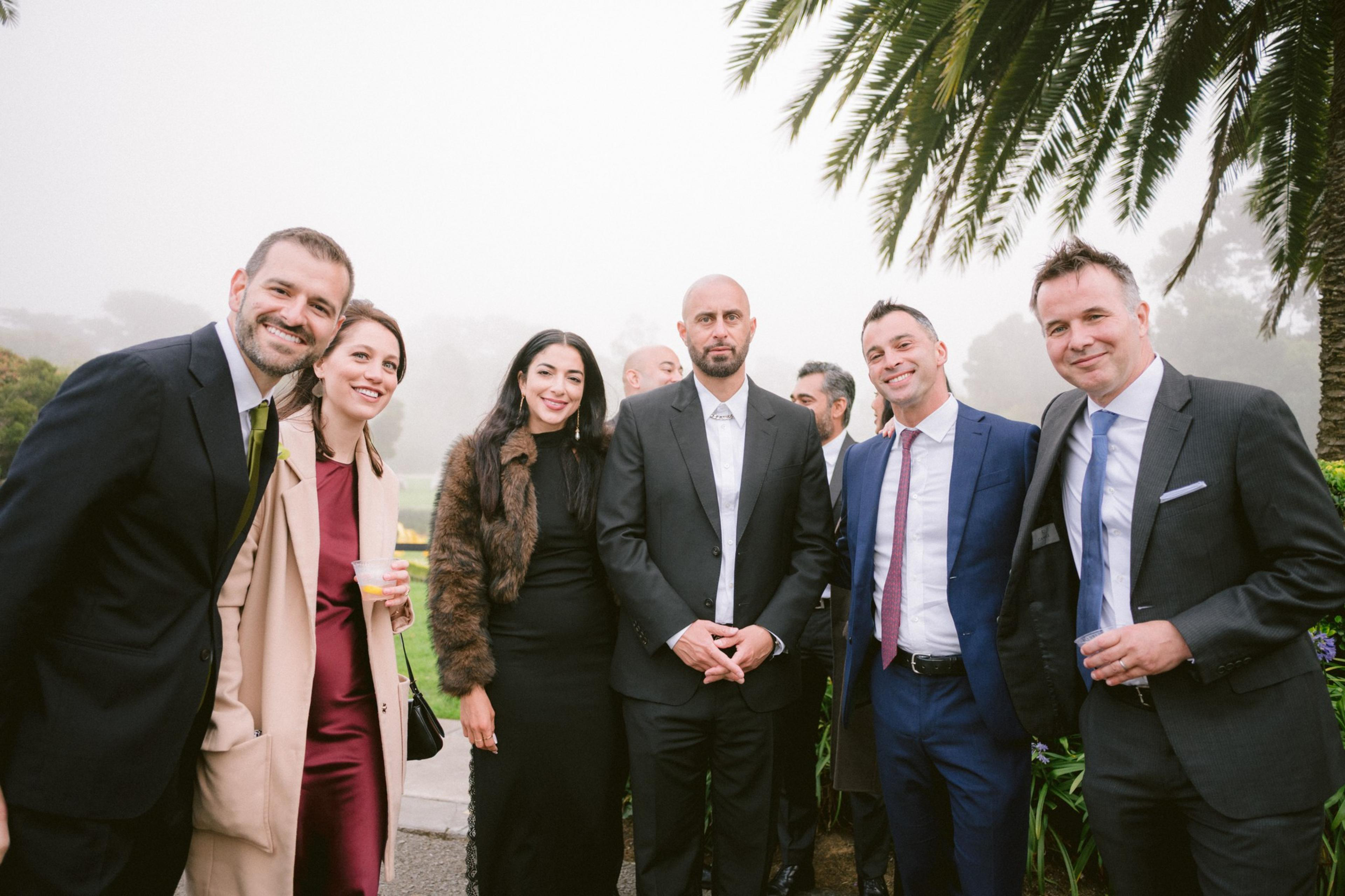 Seven people dressed in formal attire stand outdoors, smiling and posing with greenery and a palm tree visible in the foggy background.
