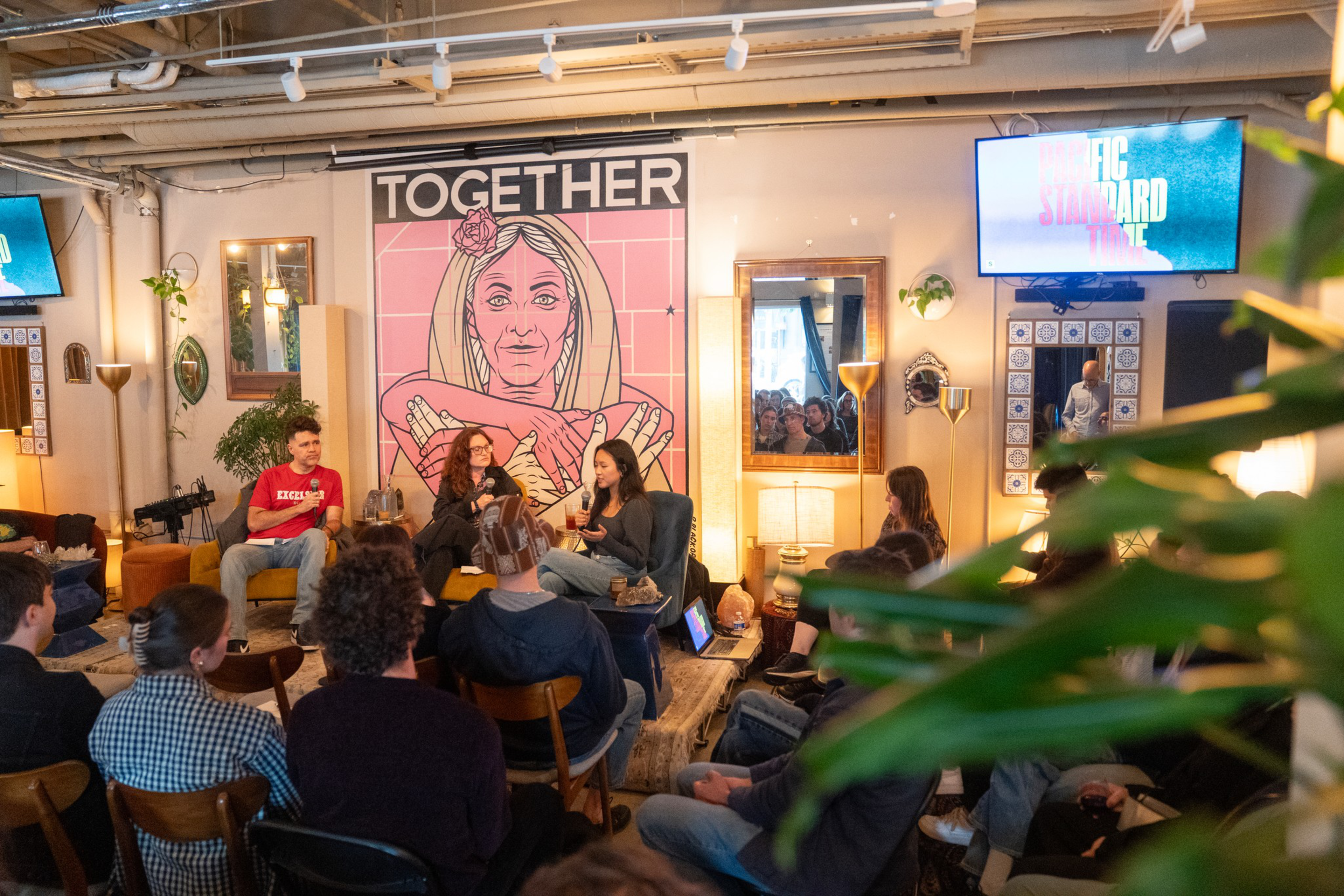A cozy room with a diverse group of people listening to three speakers seated in front of a large “TOGETHER” mural featuring a woman with folded arms.