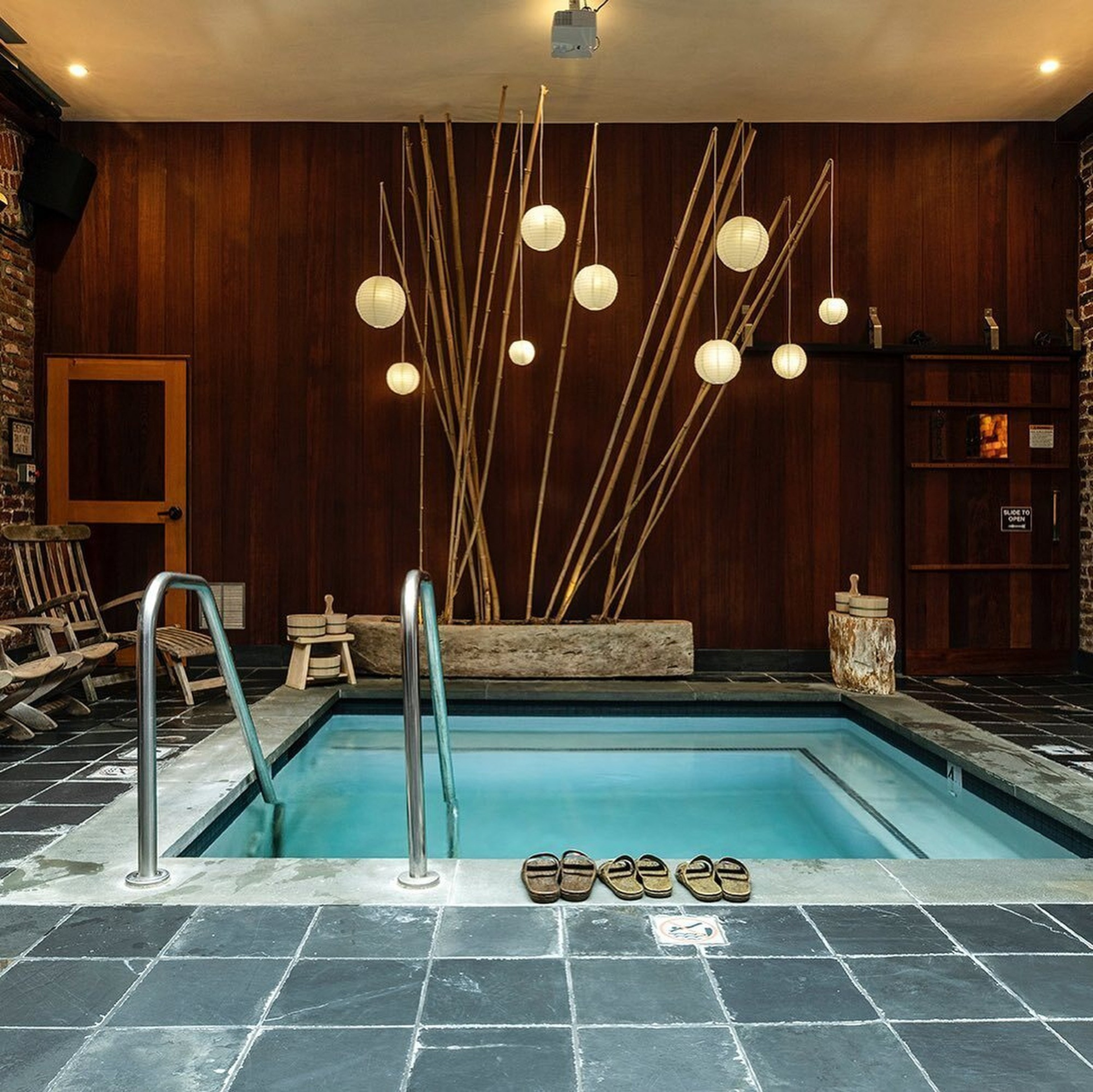 The image shows an indoor pool with glowing orb lights hanging above bamboo sticks. Wooden decor and chairs surround the pool on a dark tiled floor.