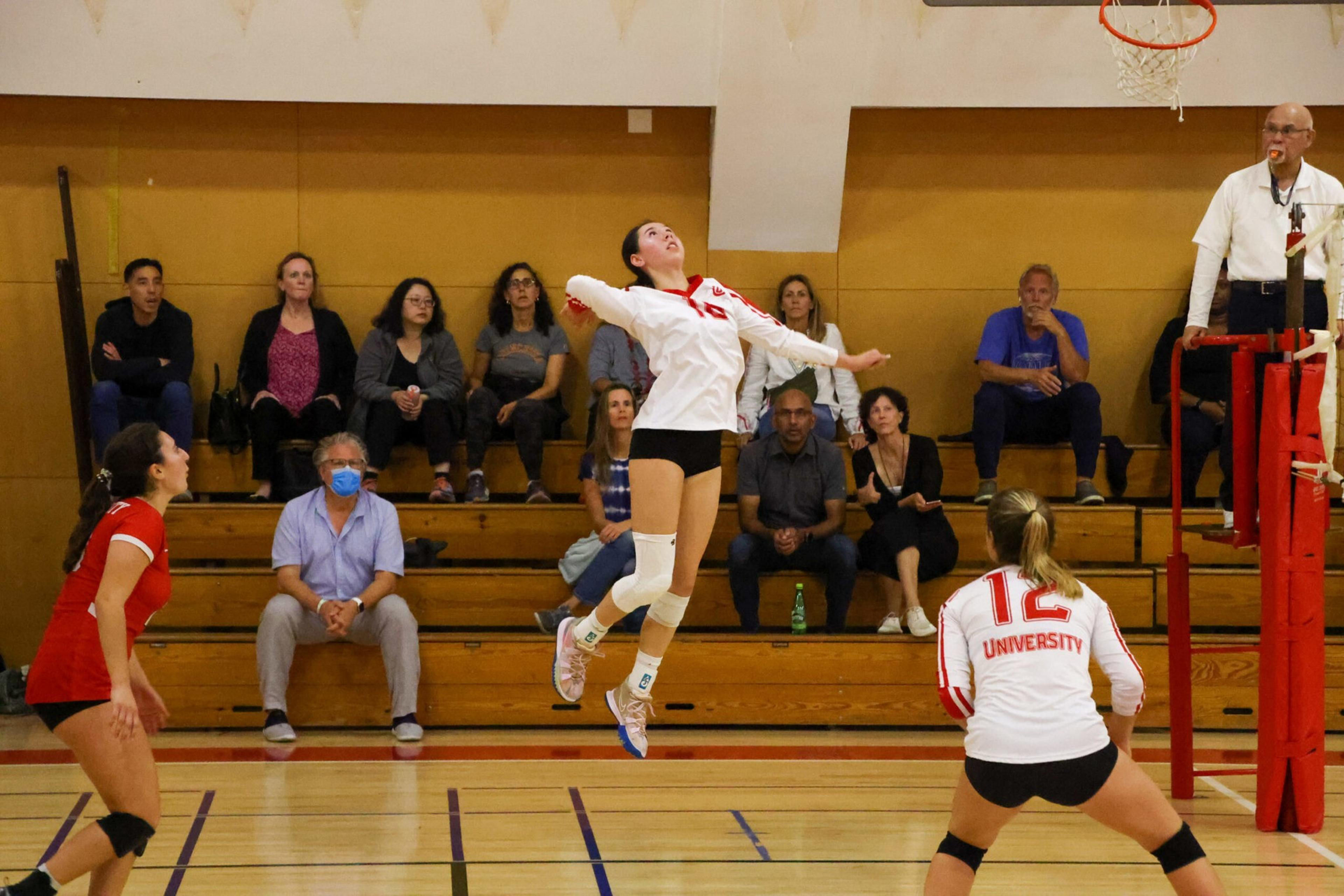 A volleyball player in a white jersey jumps to spike the ball while teammates and spectators watch from bleachers in a gymnasium.