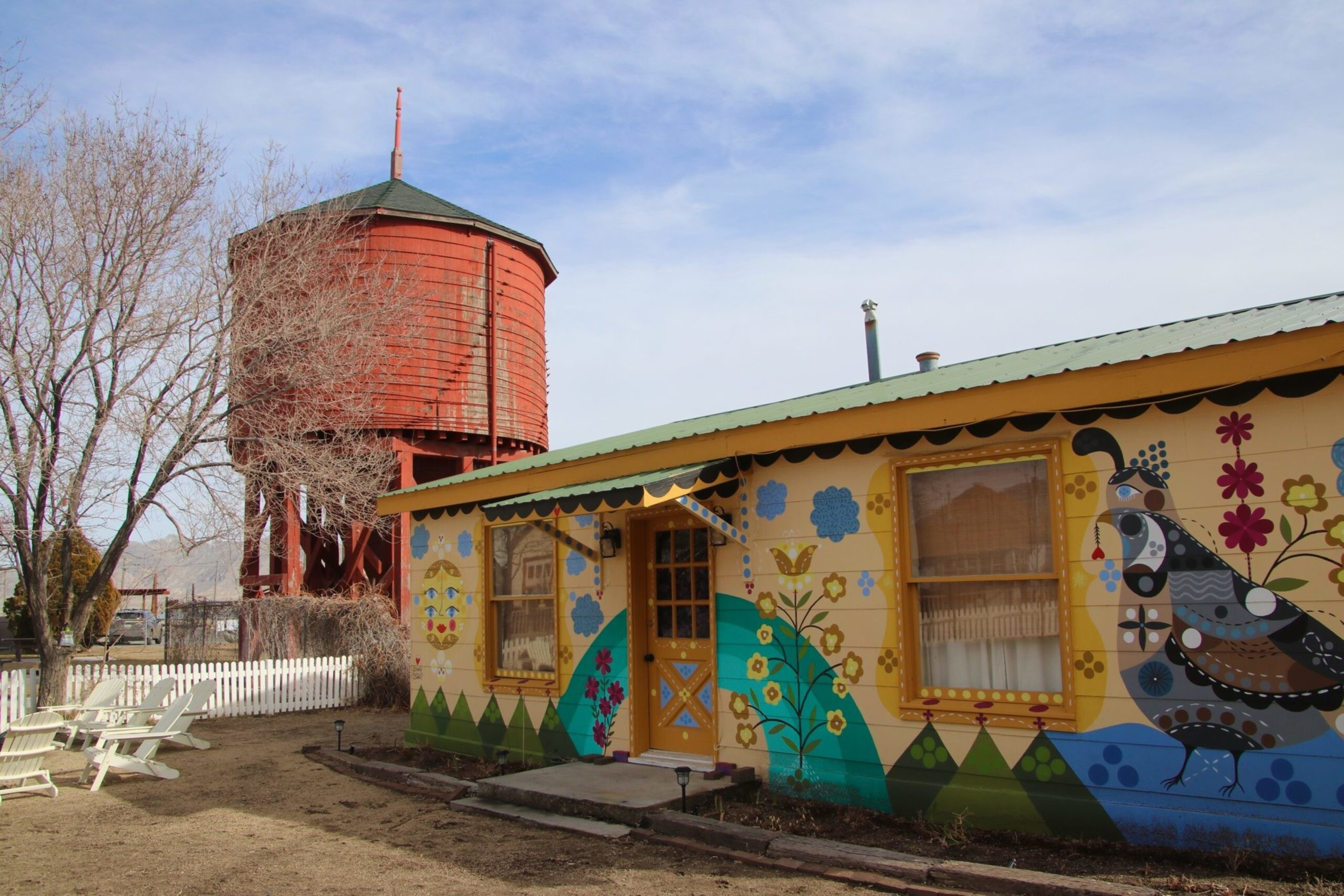 A small house painted with colorful flowers, birds, and butterflies sits beside a tall red wooden water tower and leafless trees.