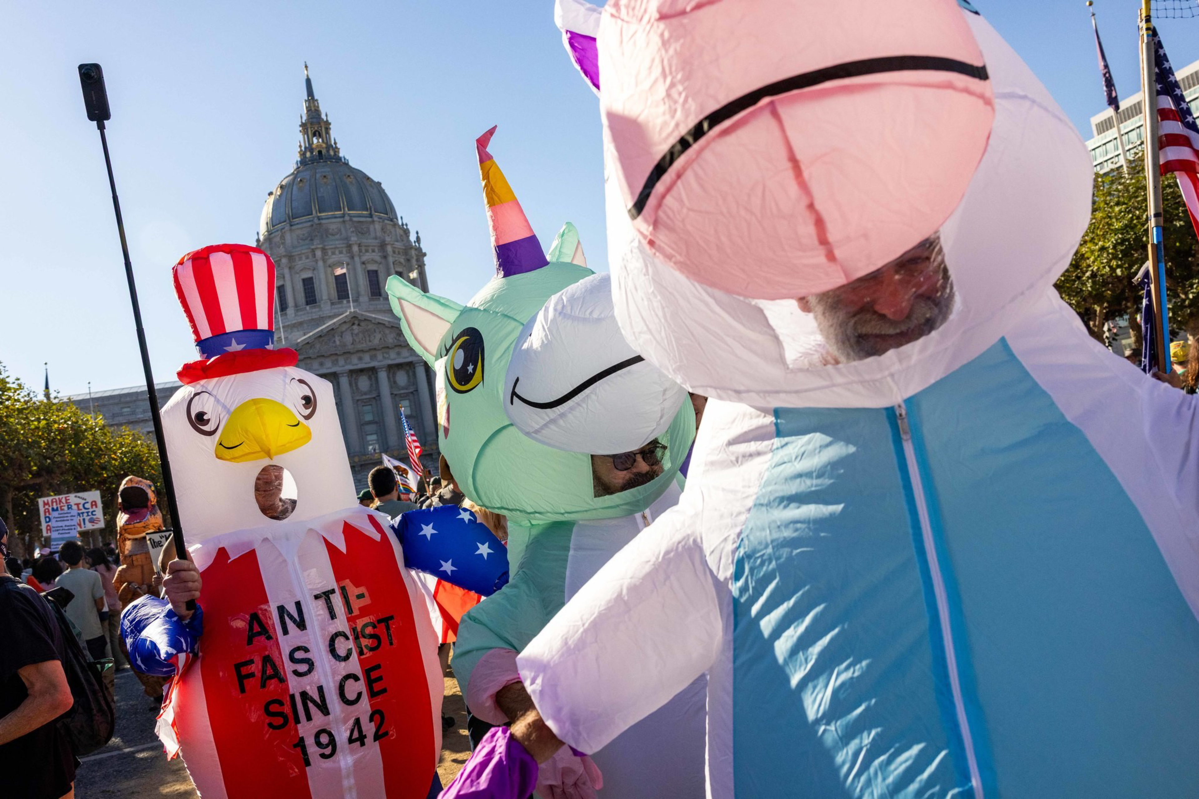 People wearing large inflatable costumes of a unicorn, a dinosaur, and an eagle with a sign reading “ANTI-FASCIST SINCE 1942" at a public event.