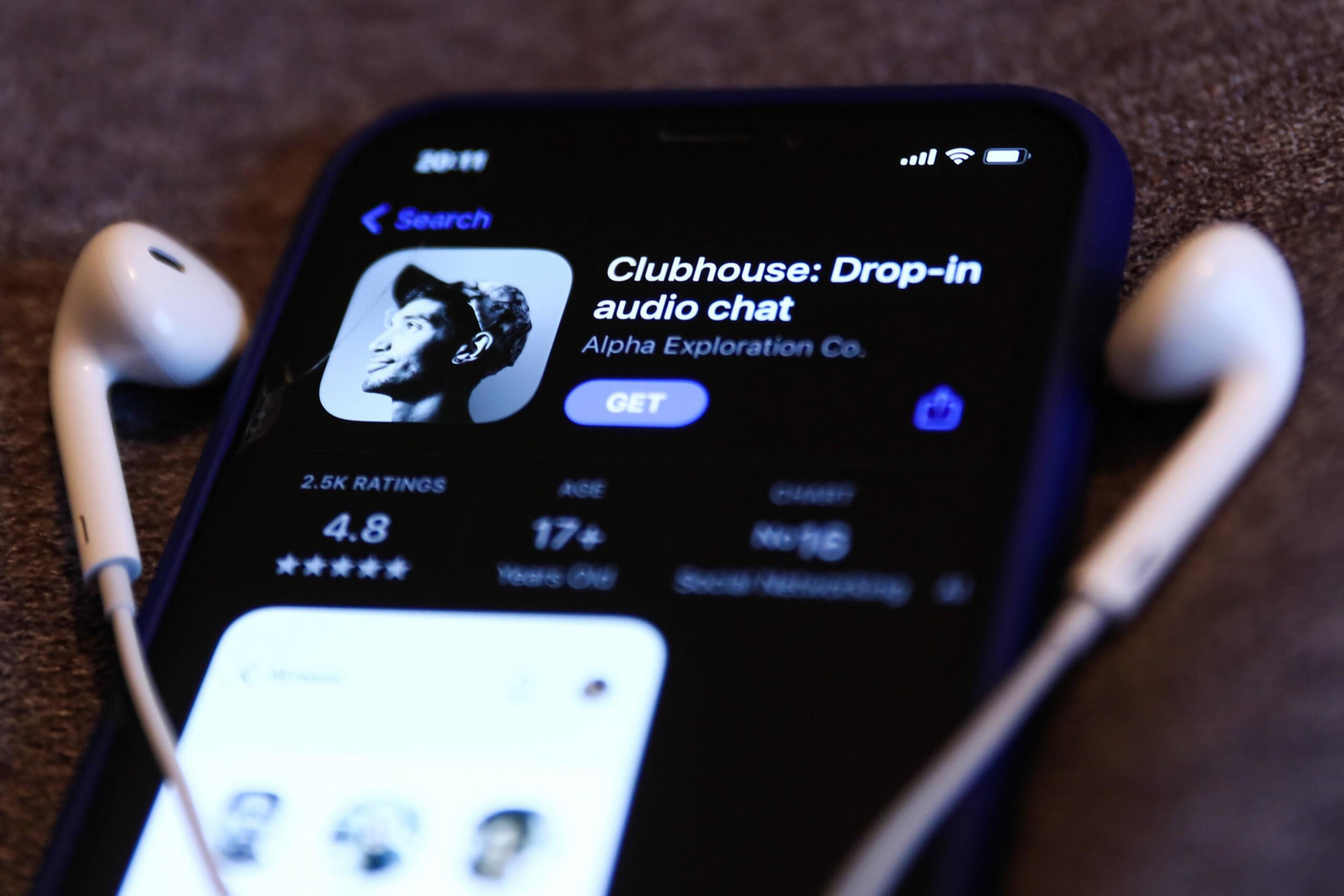 A phone screen displays the Clubhouse app page with white earphones placed on either side of the phone.
