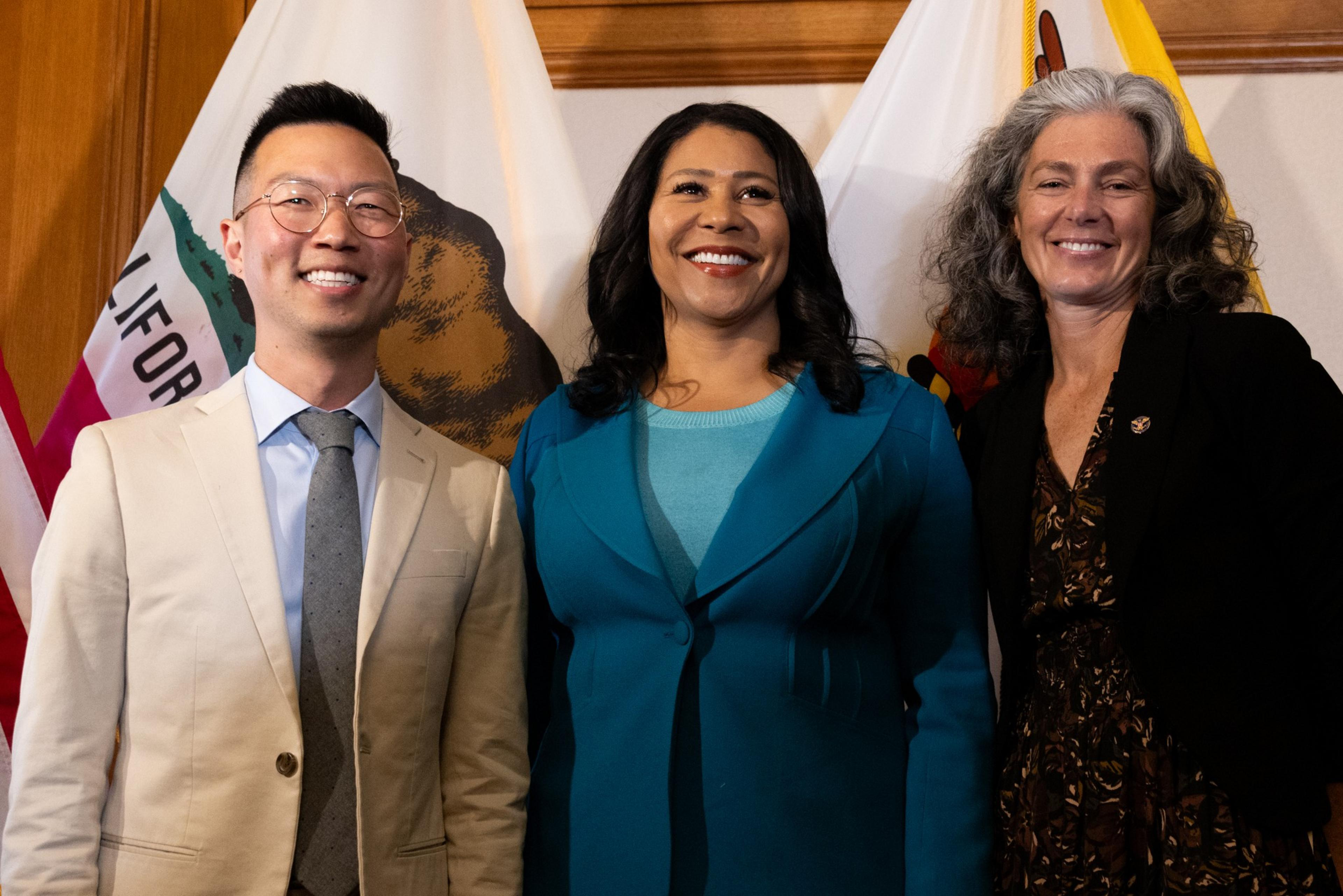 Three people are posing and smiling in front of California flags: a man in a beige suit, a woman in a turquoise jacket, and a woman in a black blazer.