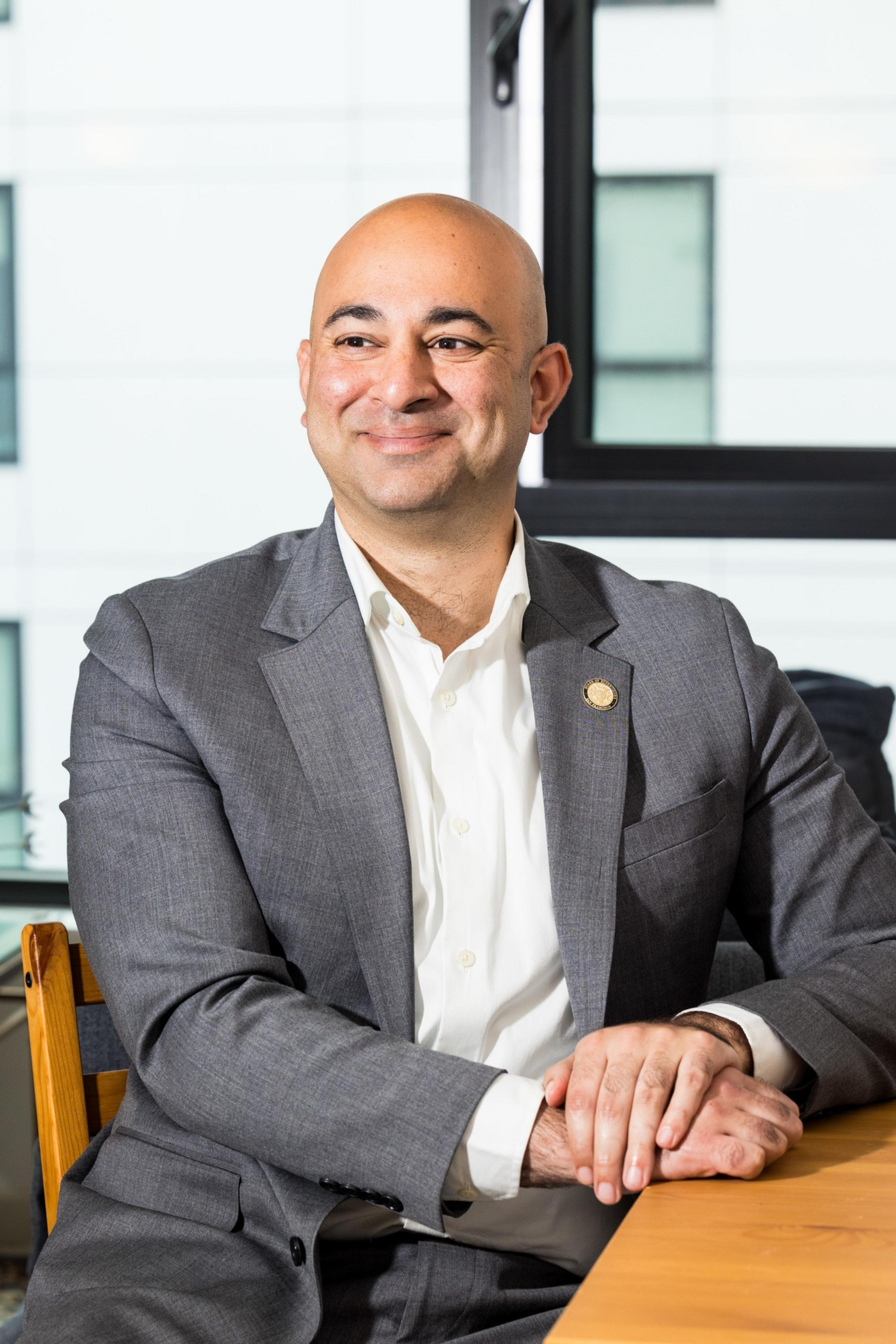 A smiling man in a gray suit and white shirt sits at a table with his hands crossed, in front of a large window with a blurred modern building background.