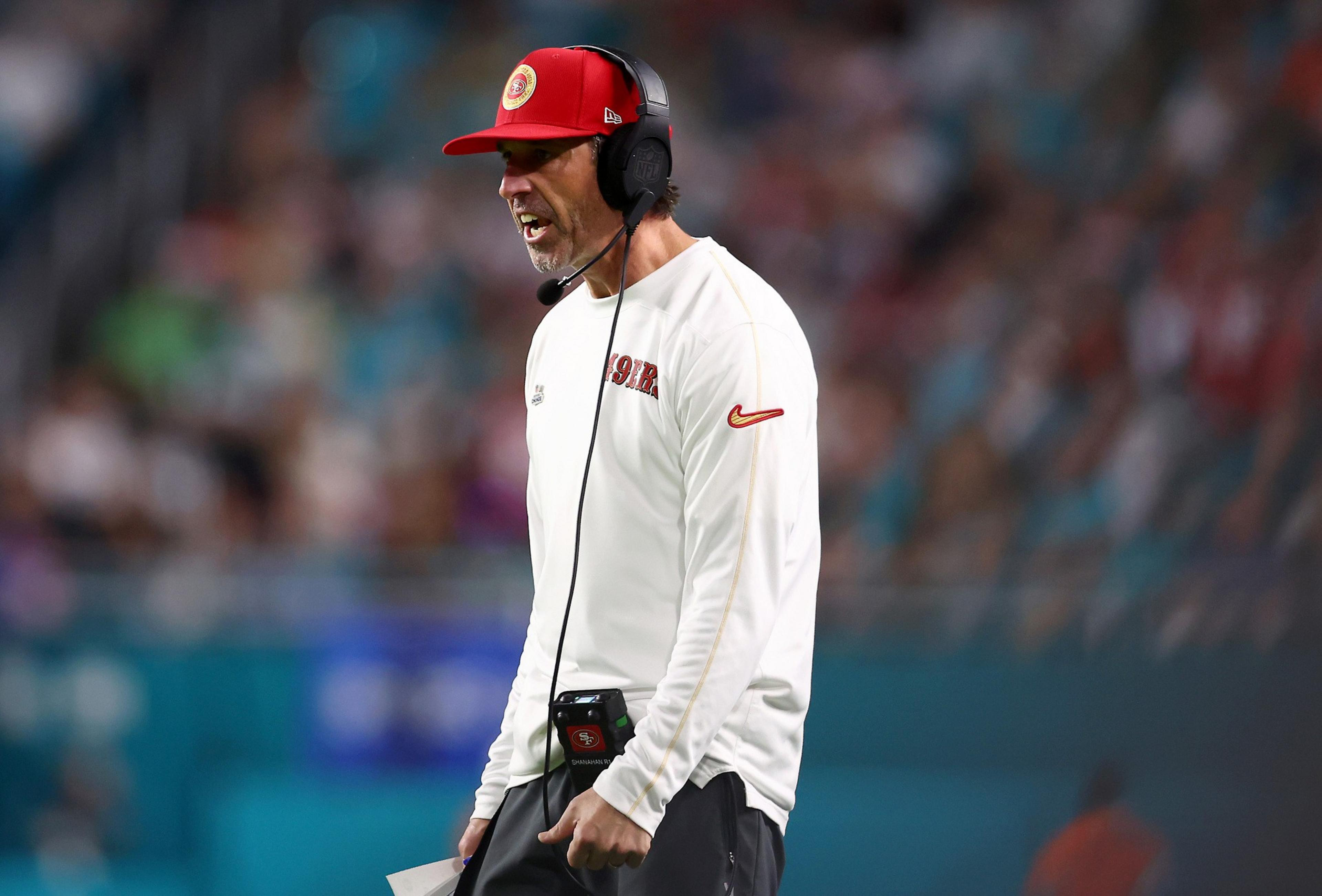 A man on a football field wears a red cap and headphones. Dressed in a white long-sleeve shirt with "49ers" on it, he appears to be shouting instructions.