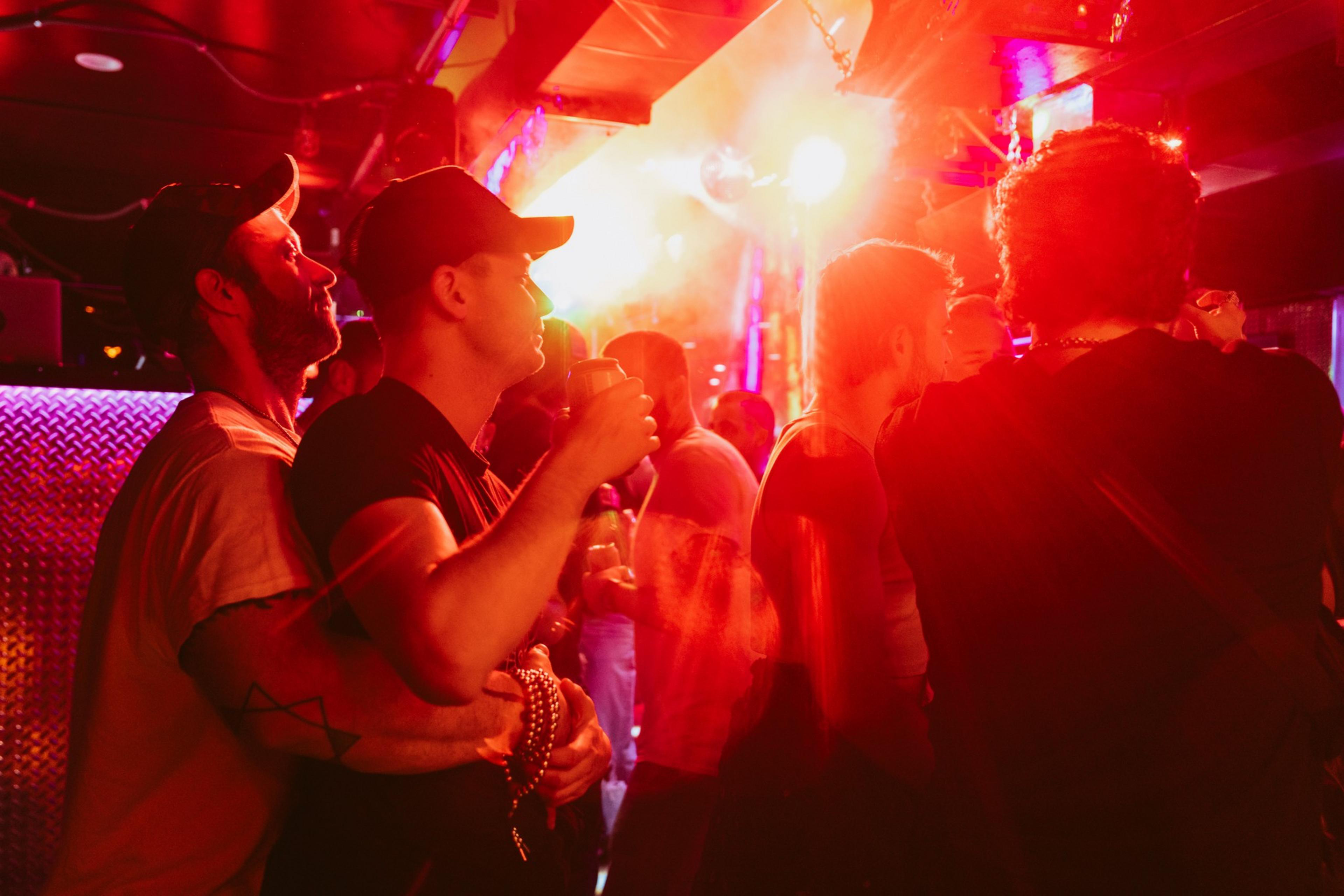 A group of people stands in a dimly lit nightclub with red and pink lighting, creating a warm atmosphere. Some are holding drinks, and there's a sense of lively conversation.