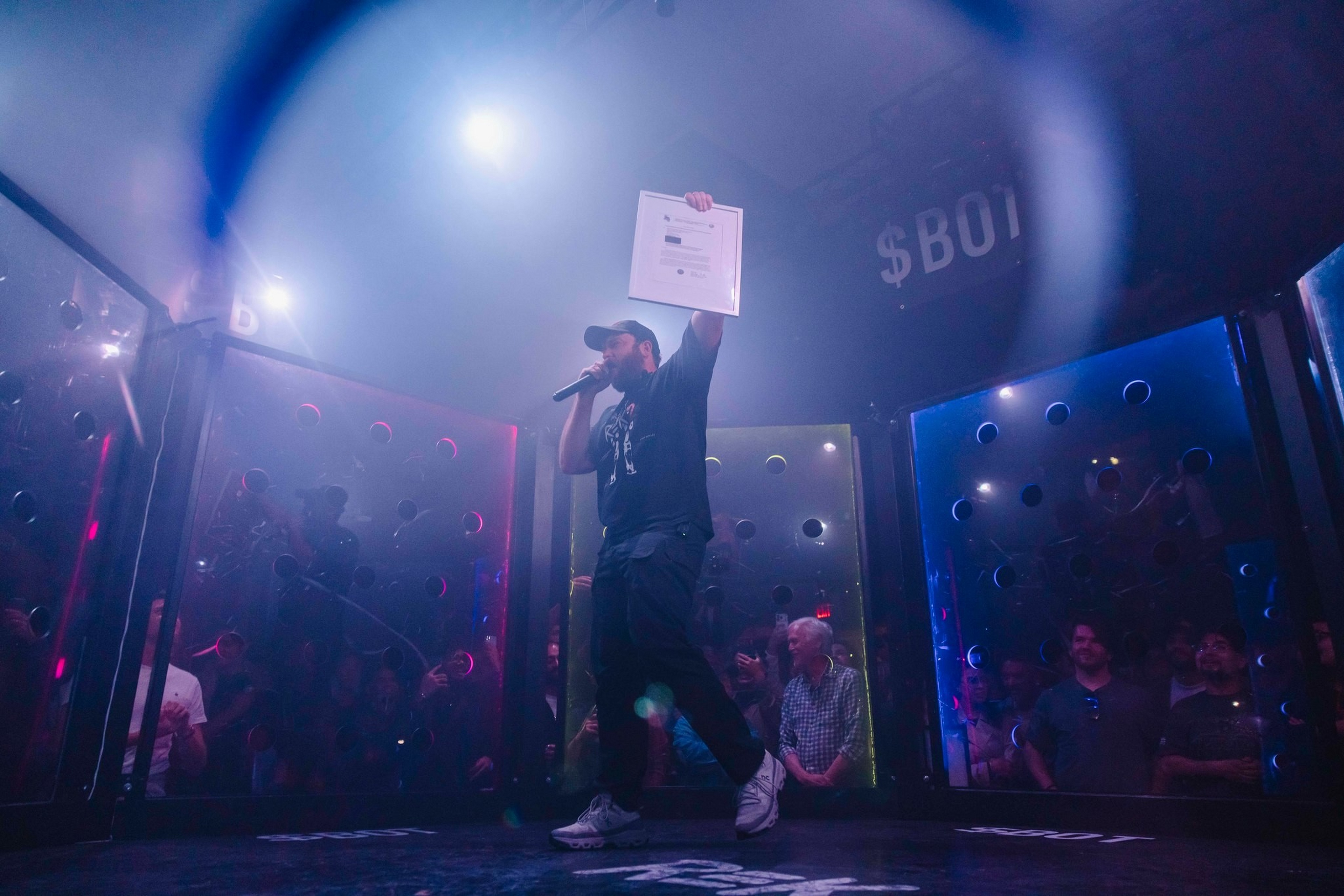 A man wearing a cap holds a microphone and raises a framed certificate on stage, surrounded by an audience behind transparent panels in a dim venue.
