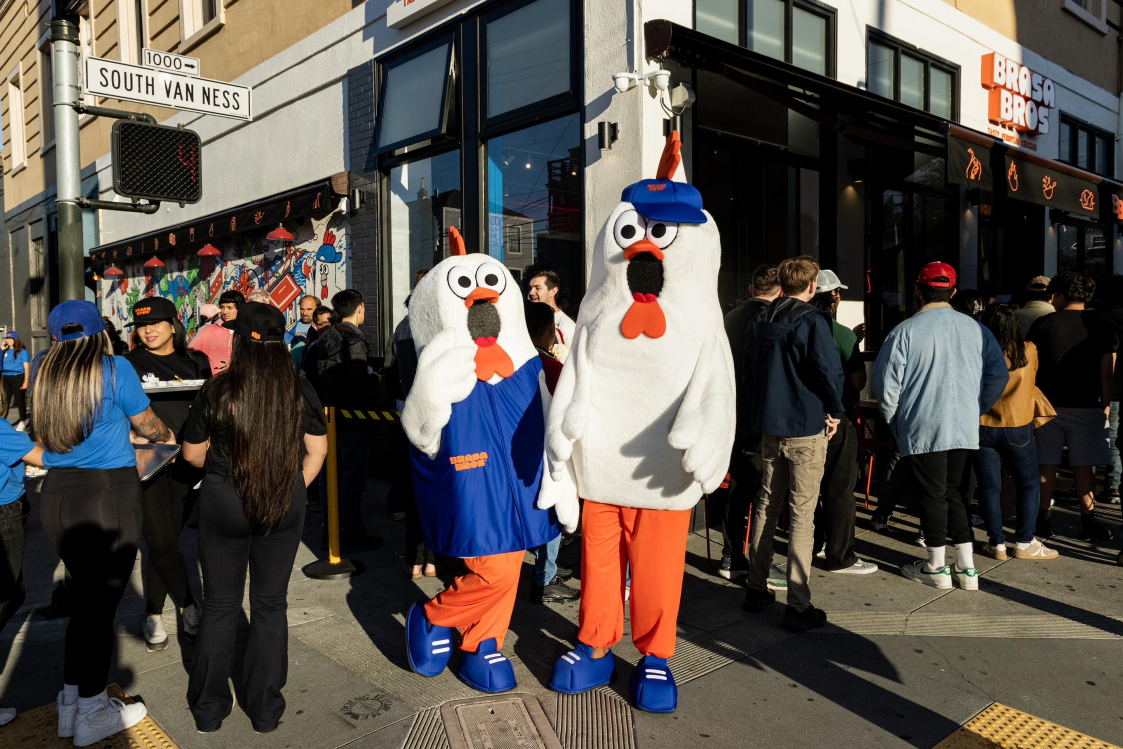 Two people in chicken costumes with blue and orange outfits stand outside a crowded restaurant on a sunny street corner at South Van Ness.