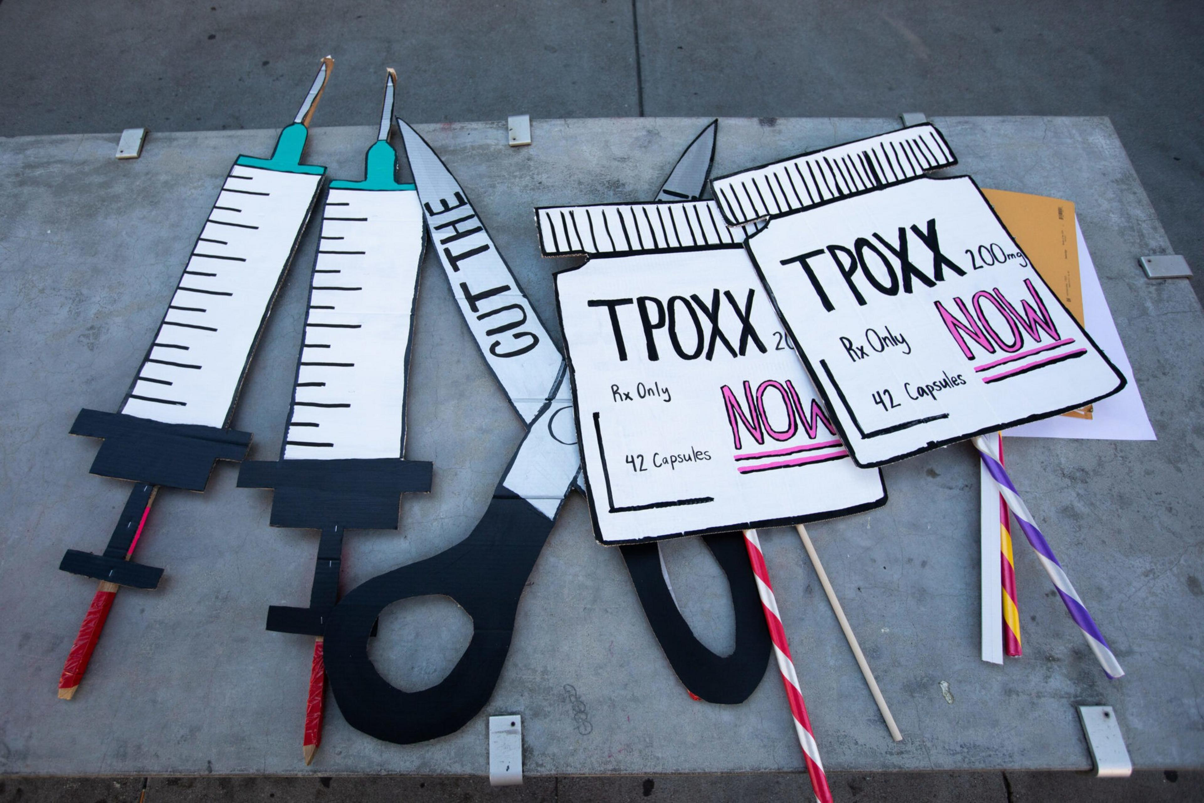 Two large syringe cutouts, a pair of scissors labeled "Cut the," and two oversized pill bottle cutouts labeled "TPOXX NOW" lie on a concrete surface.