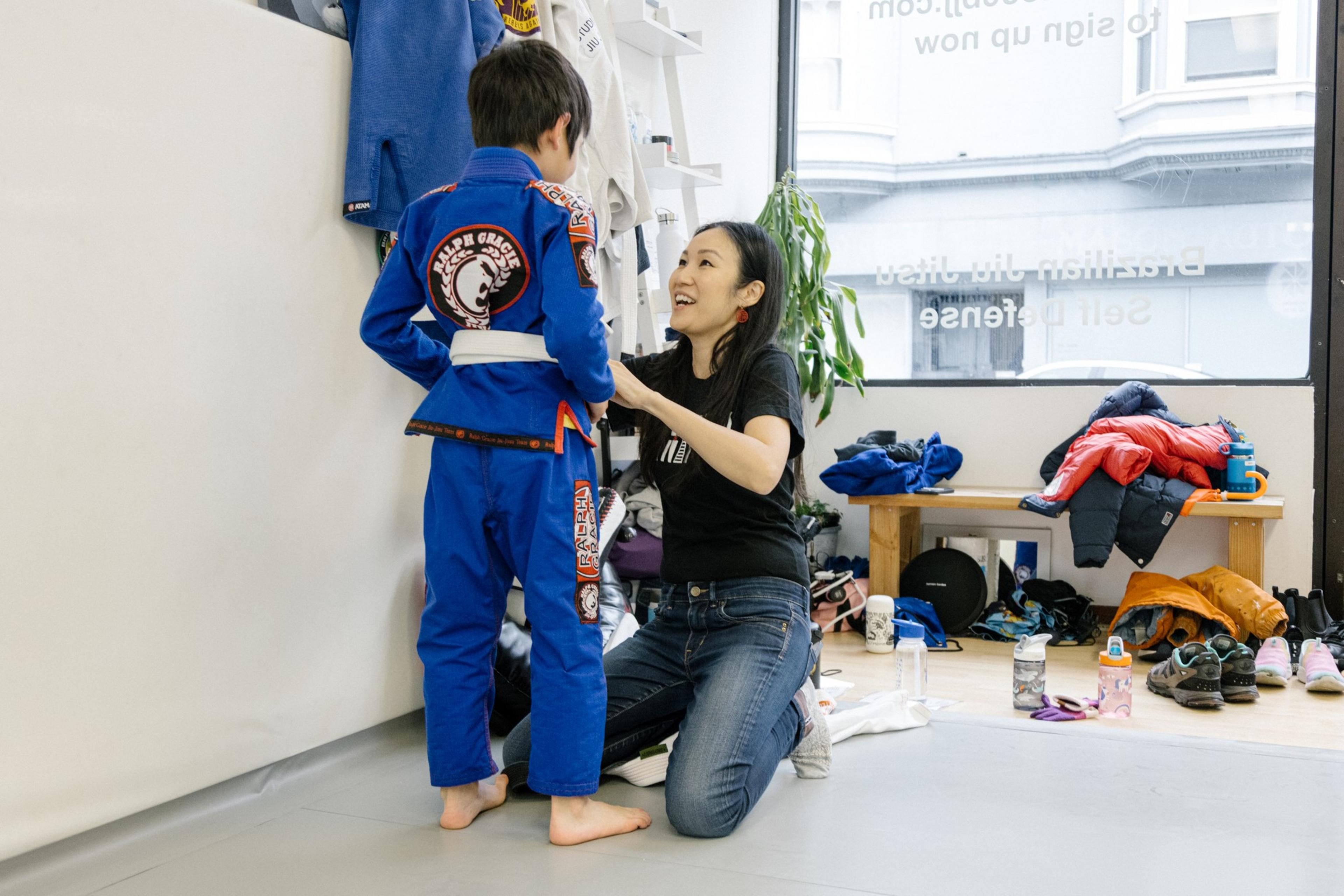 A woman is kneeling, tying the belt of a child in a blue judo uniform, both smiling in a dojo with gear on shelves.