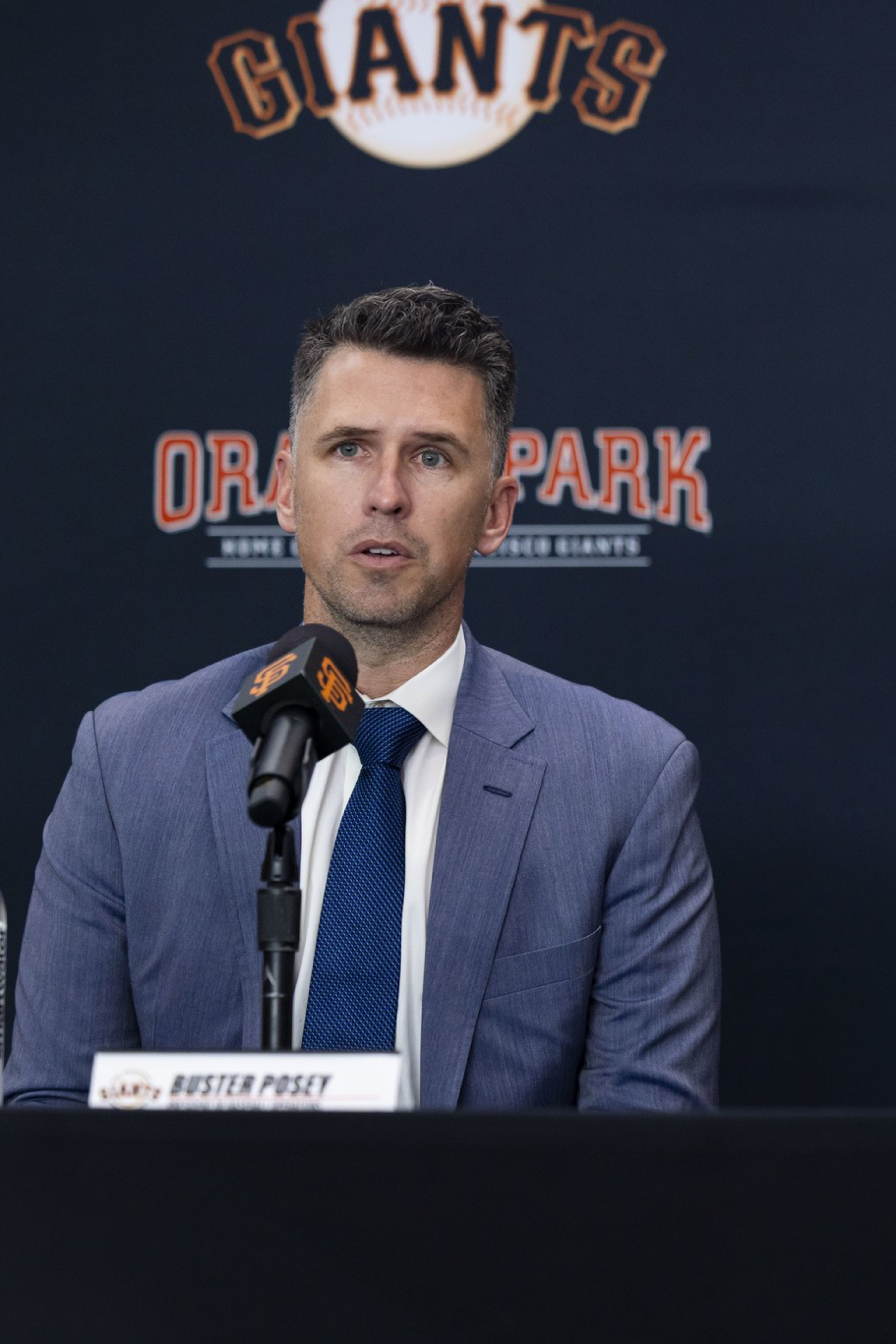 Two men sit at a press conference table with microphones, one in a suit and the other in a San Francisco Giants uniform and cap.