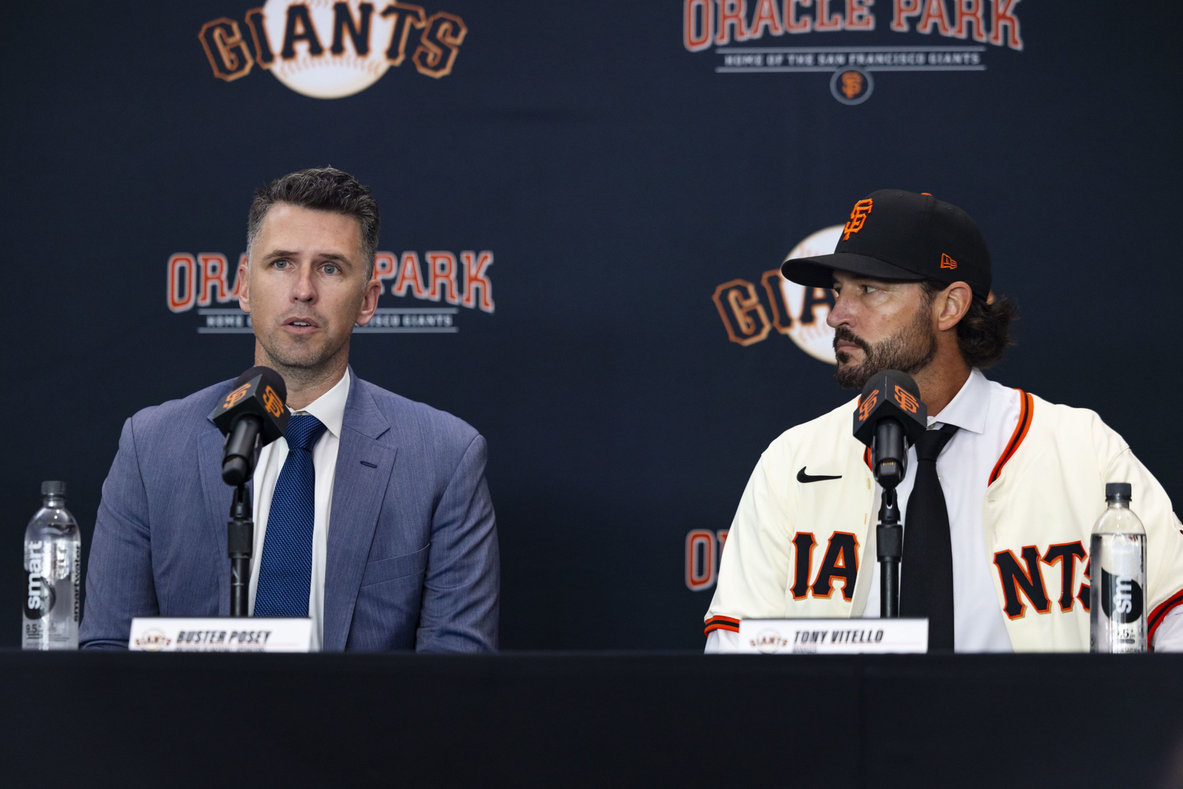 Two men sit at a press conference table with microphones, one in a suit and the other in a San Francisco Giants uniform and cap.