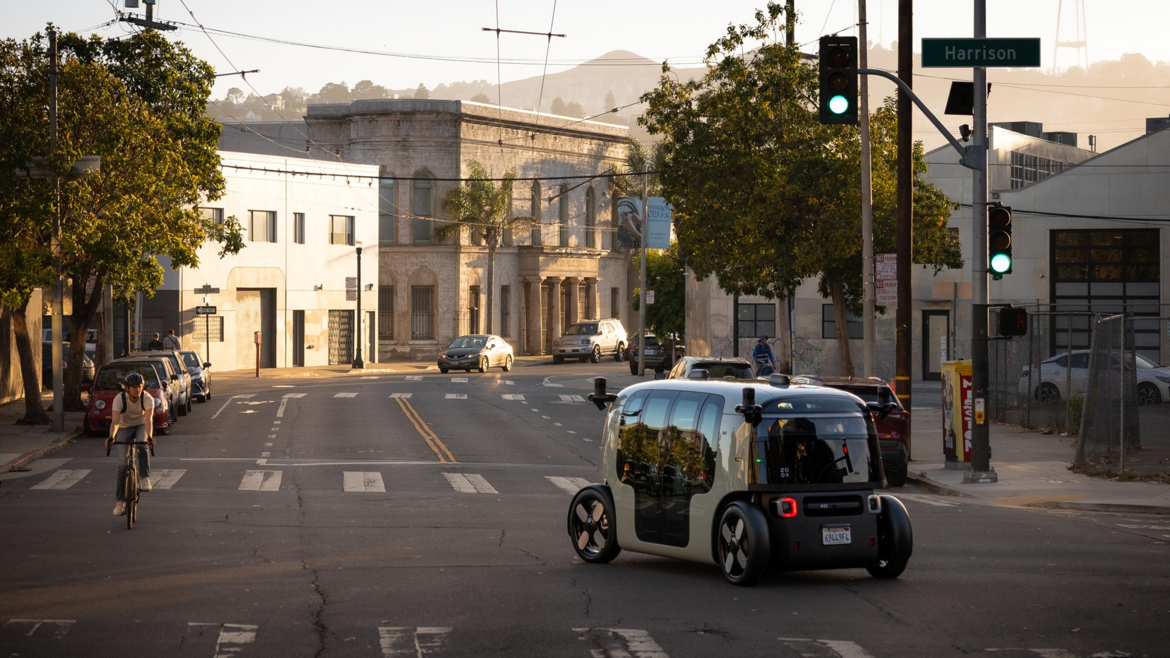 A small, futuristic autonomous vehicle drives through a sunny intersection near a Harrison street sign, with a cyclist and several parked cars nearby.