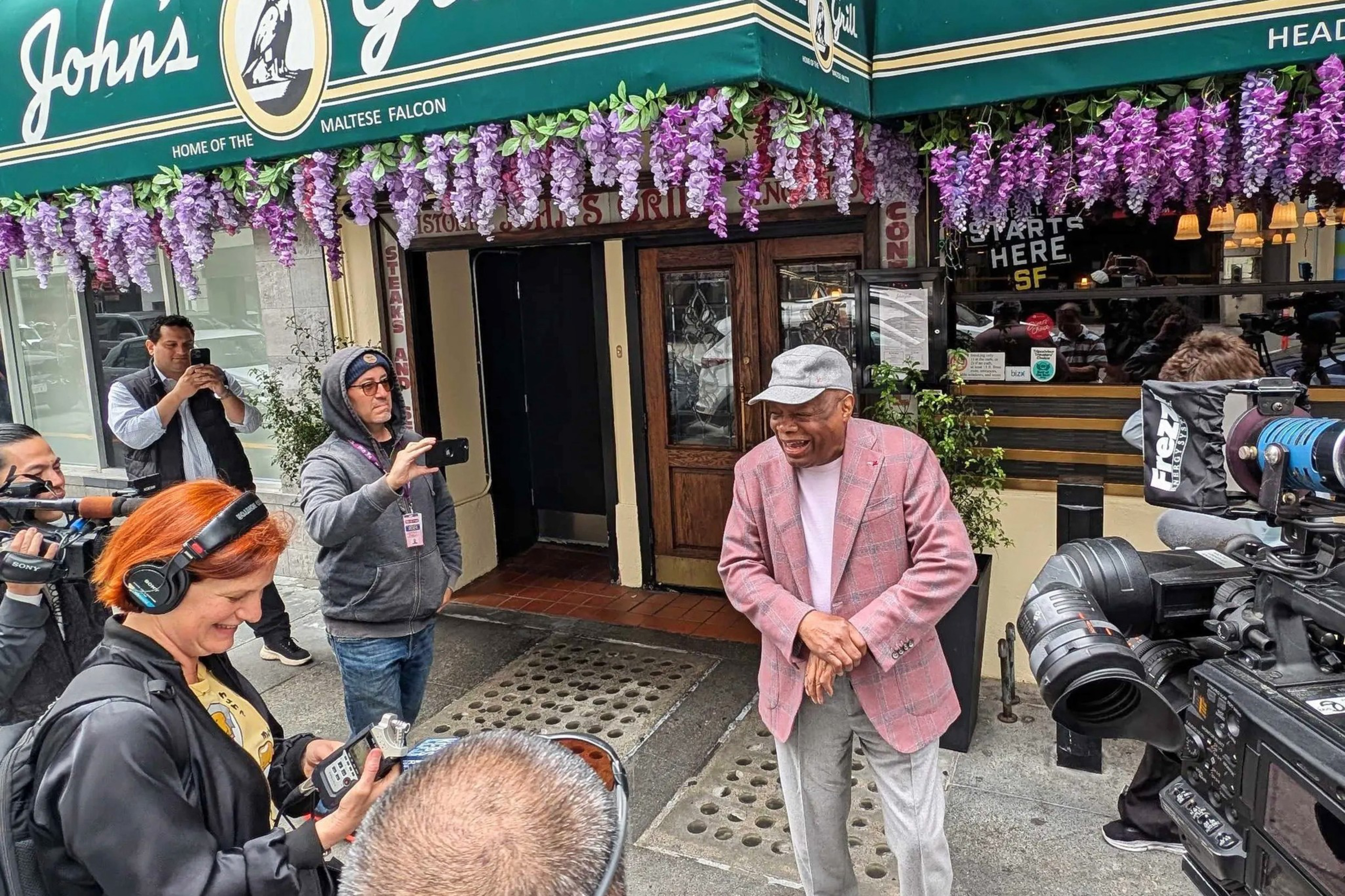 A man in a pink checked blazer and gray cap stands smiling outside John's Grill, surrounded by several people with cameras and recording equipment.