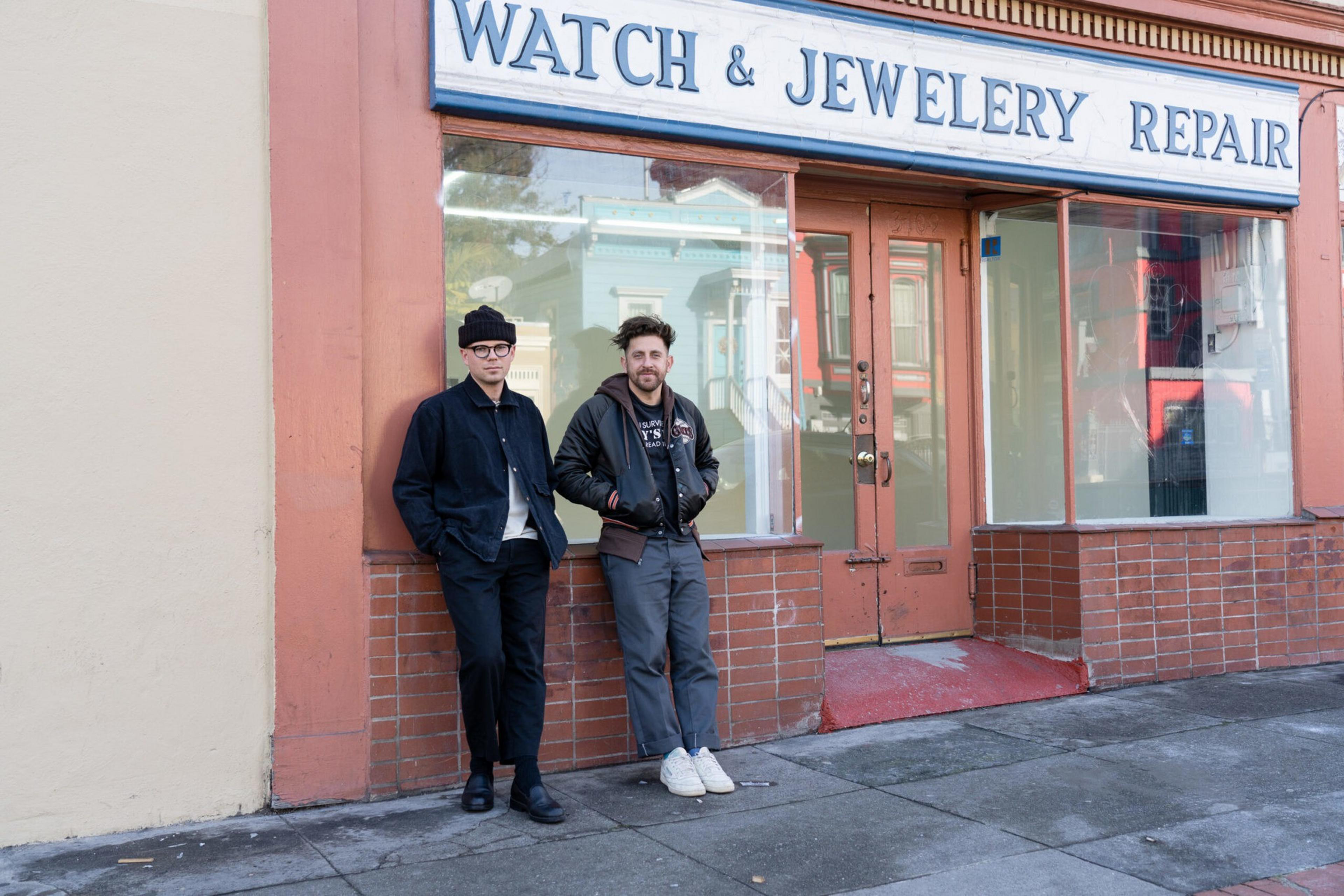 Two men stand outside a closed store with a sign reading "Watch & Jewelery Repair," leaning against the wall on a sidewalk in front of a glass door.