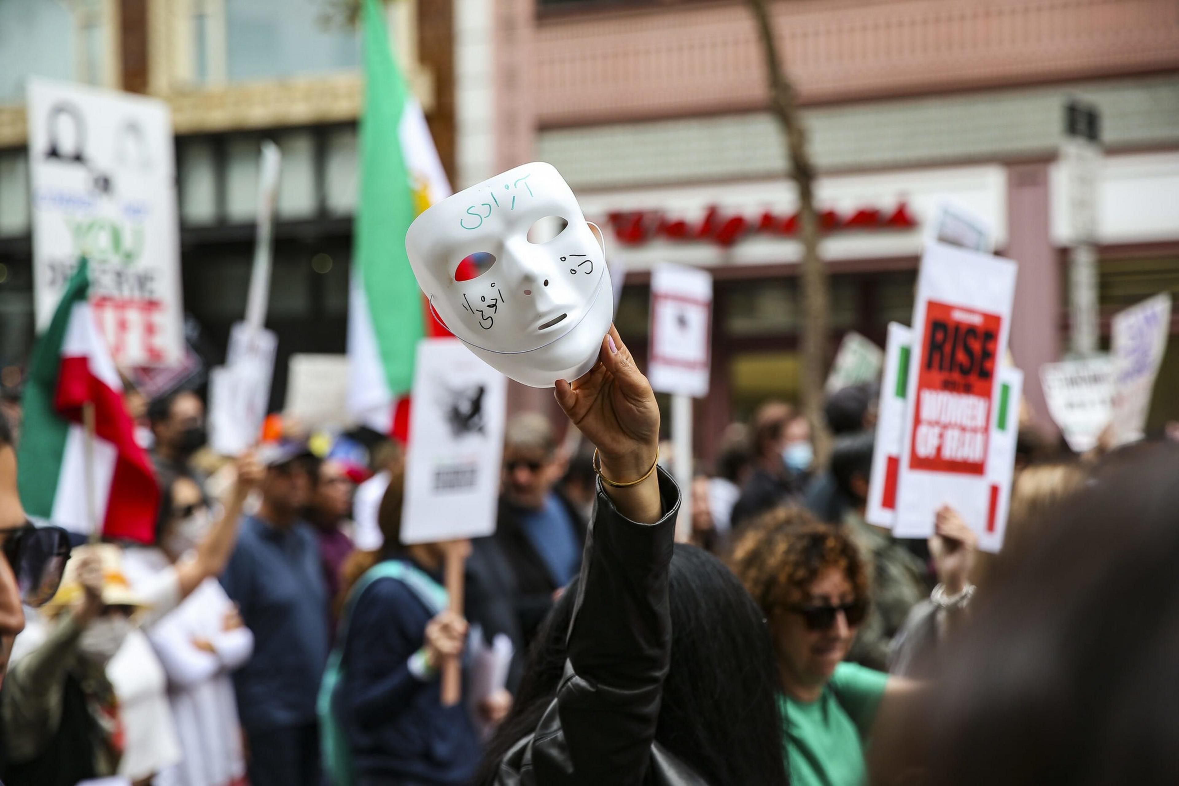 A person holds up a white mask with writing on it amid a crowd of protesters holding signs and green, white, and red flags.