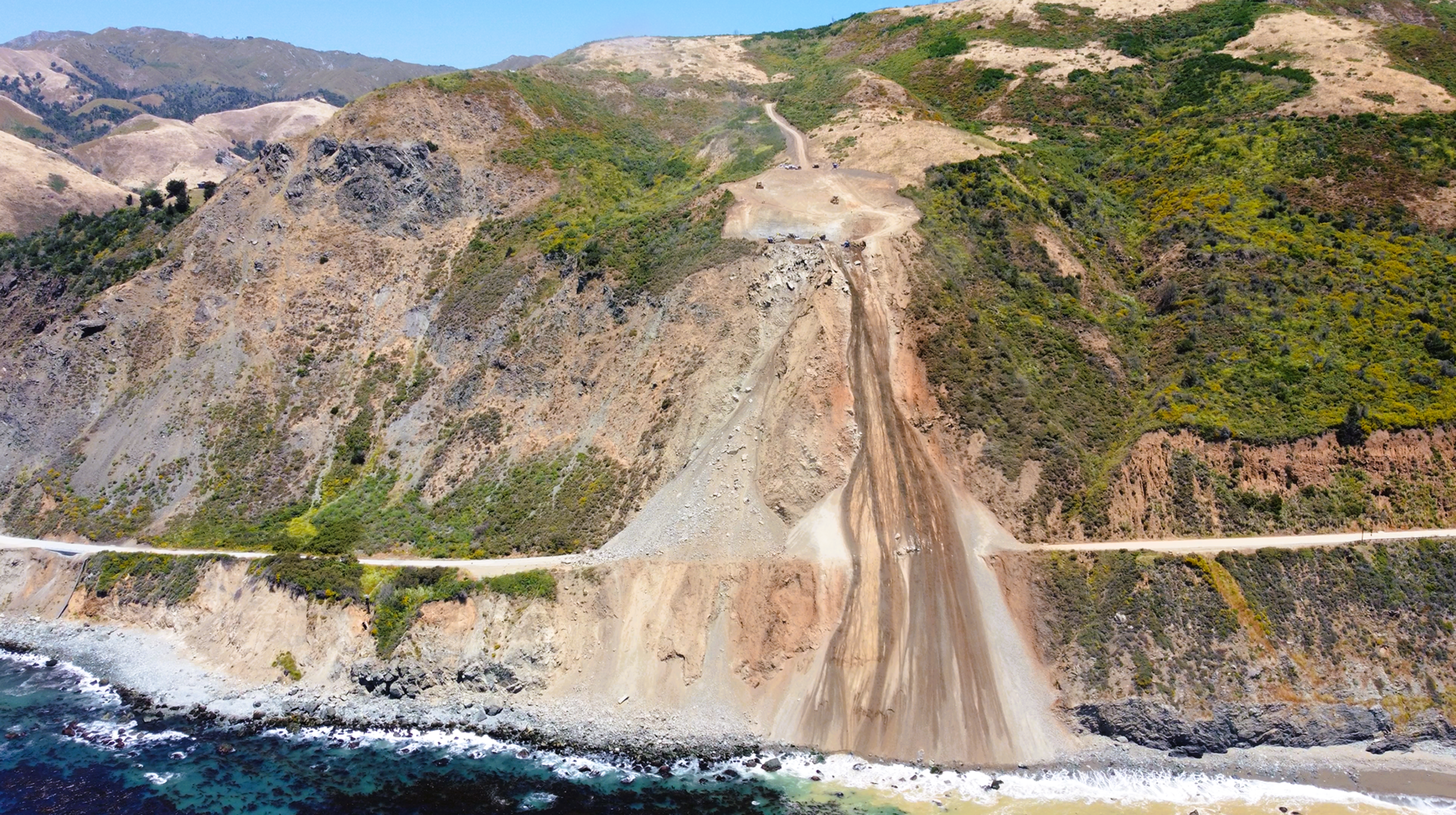 A washed-out dirt slide crosses a narrow coastal road, running down a steep, rocky hillside to a rocky beach beside a blue ocean.