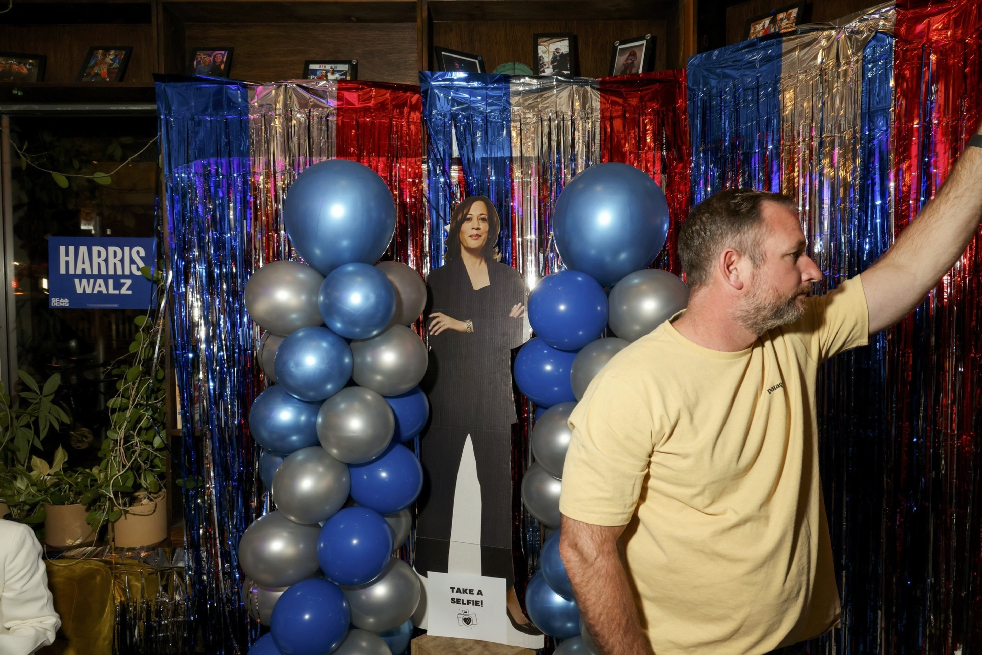 A man in a yellow shirt stands next to a cutout of a woman. Blue and silver balloons and shiny red, blue, and silver streamers decorate the background. A sign says &quot;Harris Walz.&quot;