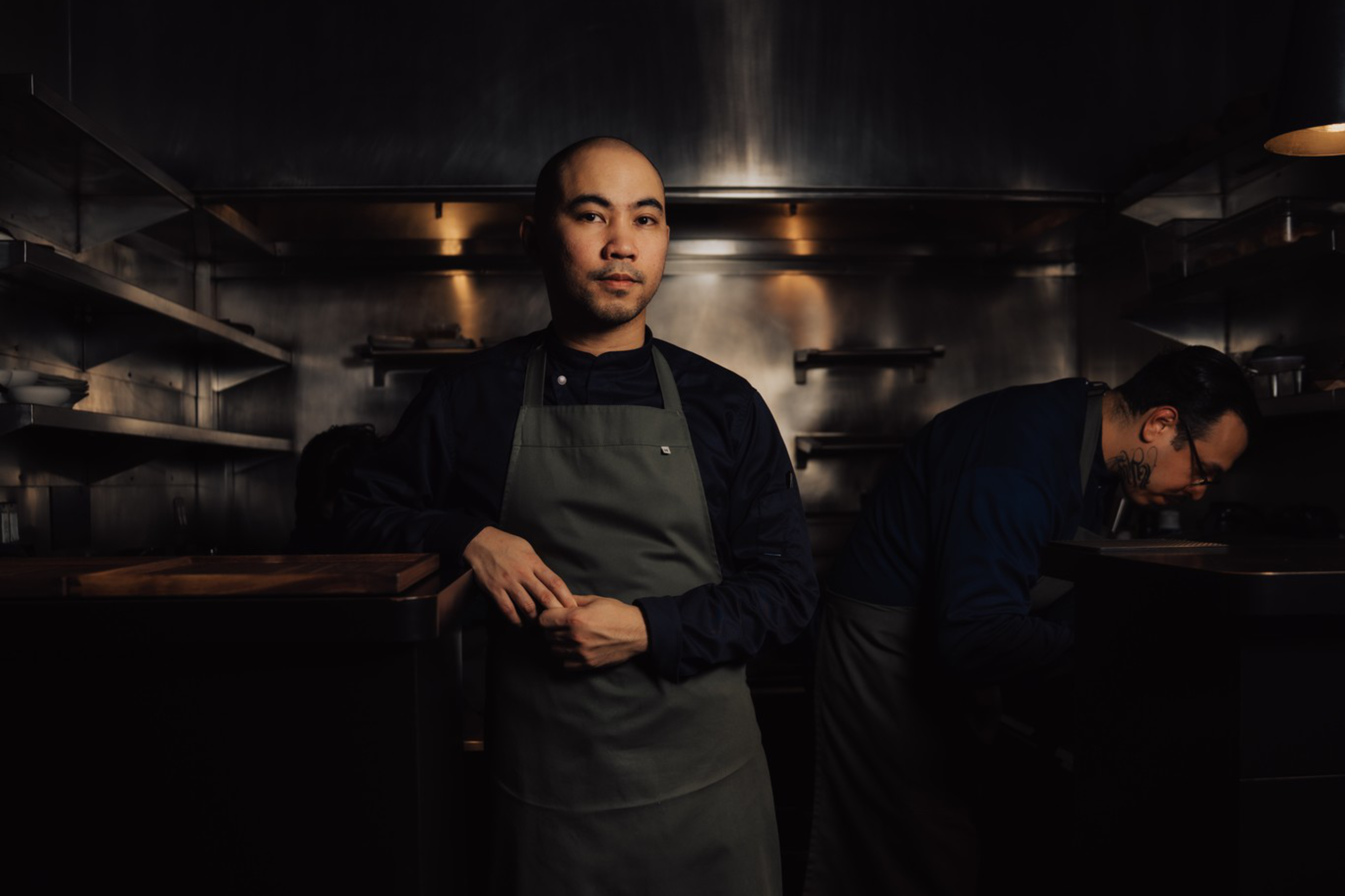 A man in a dark kitchen wears a gray apron over black clothes, standing with hands clasped, while another person works bent over in the background.