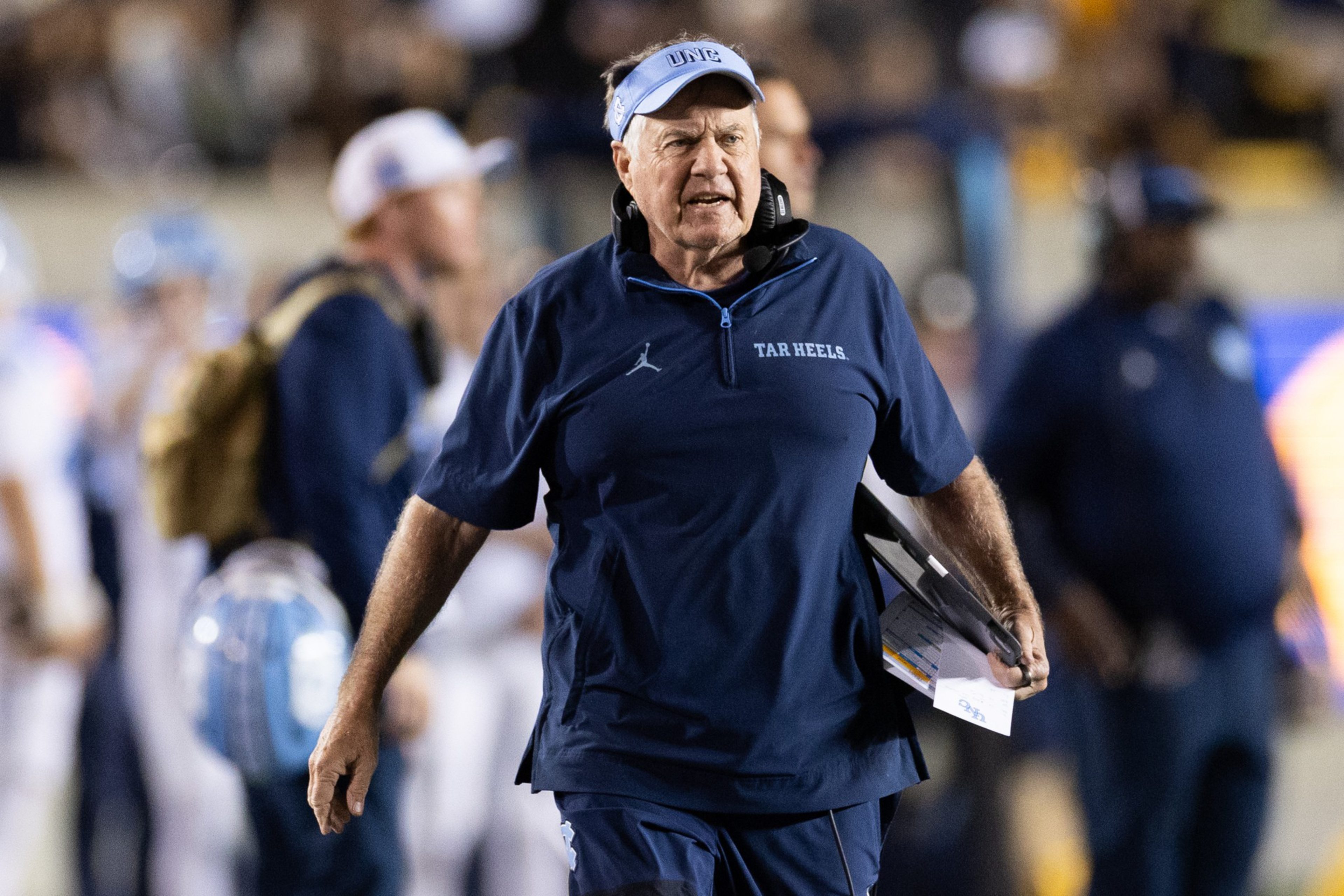A man wearing a navy blue “Tar Heels” jacket and light blue visor walks with a tablet and papers in hand on a football field.