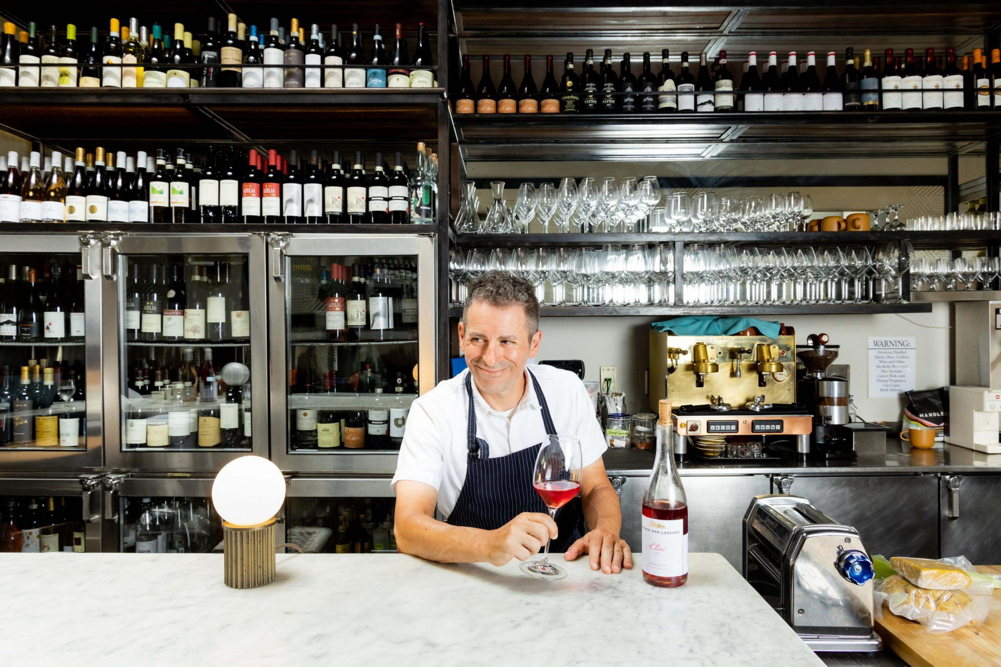 A person in a white shirt and apron stands behind a bar with wine bottles, holding a red wine glass. There are shelves with more wine bottles and glasses.