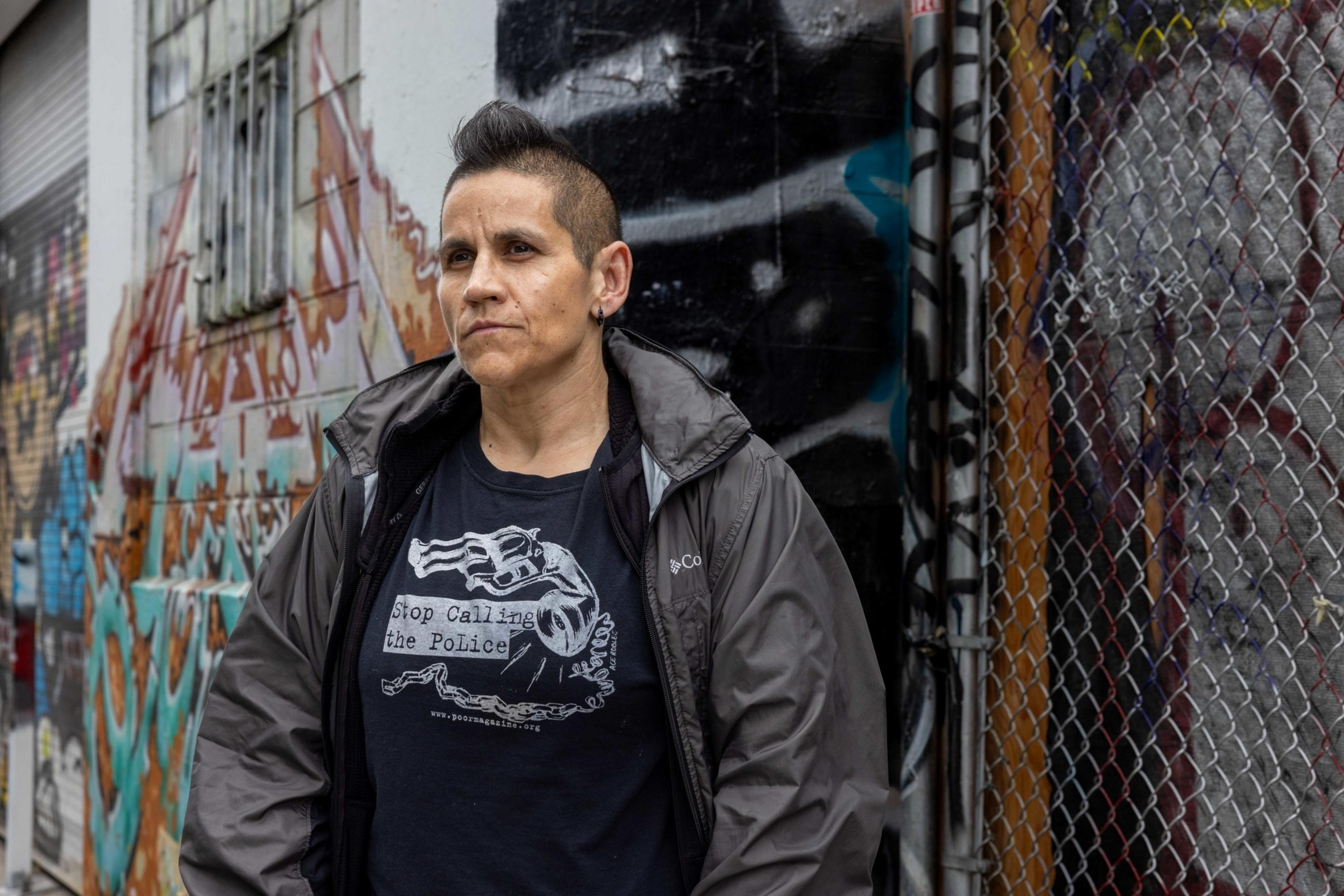 A person with short, dark hair stands in front of a graffiti-covered wall and a chain-link fence, wearing a dark jacket and a shirt that reads, &quot;Stop Calling the Police.&quot;
